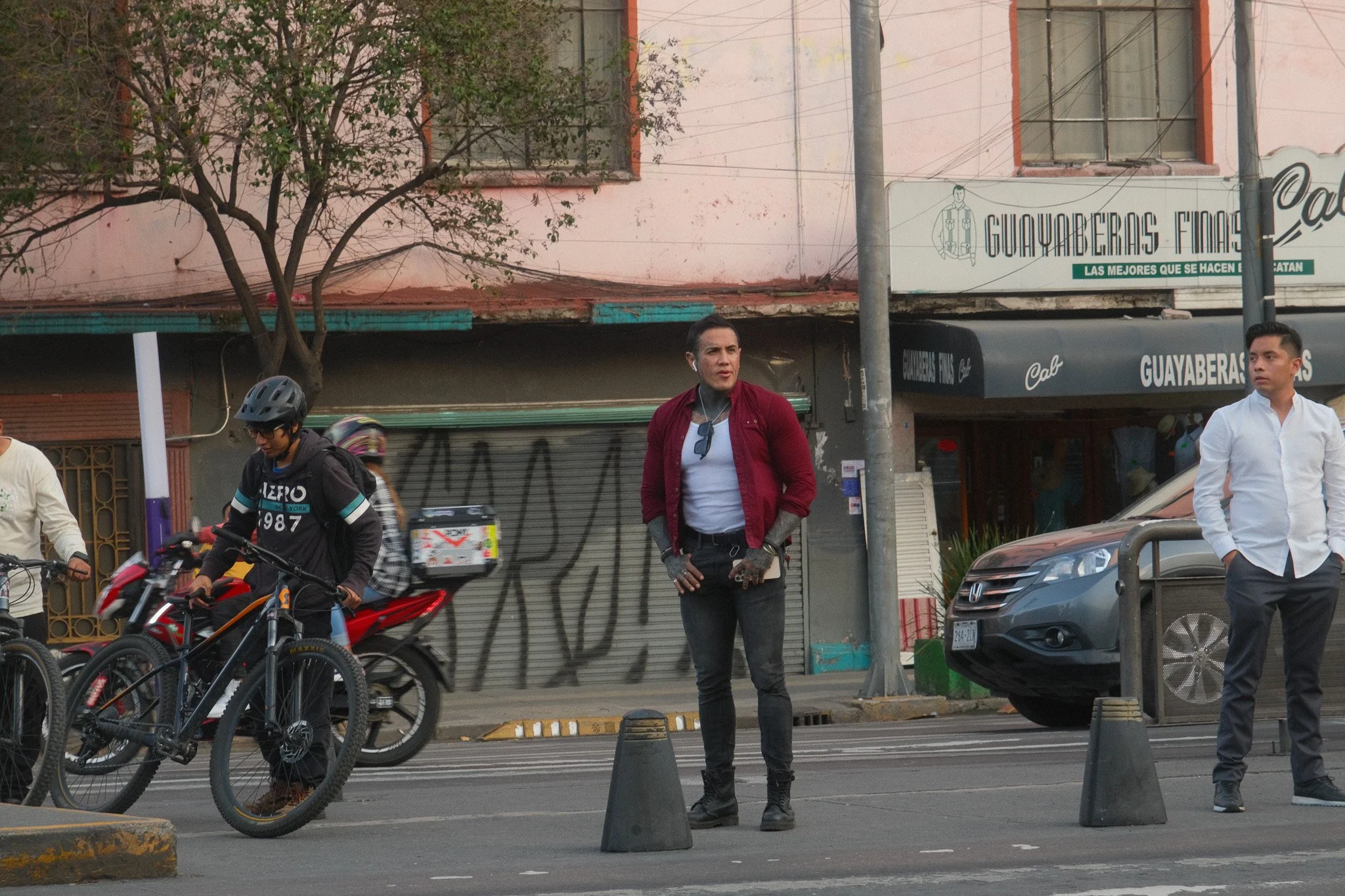 Three people standing on the sidewalk of a street with motorcycles and cars passing by. One woman in a red jacket, black pants, and tattoos on her hands, standing with her hands on her hips. A man in a white shirt with his hands in his pockets. Anoth