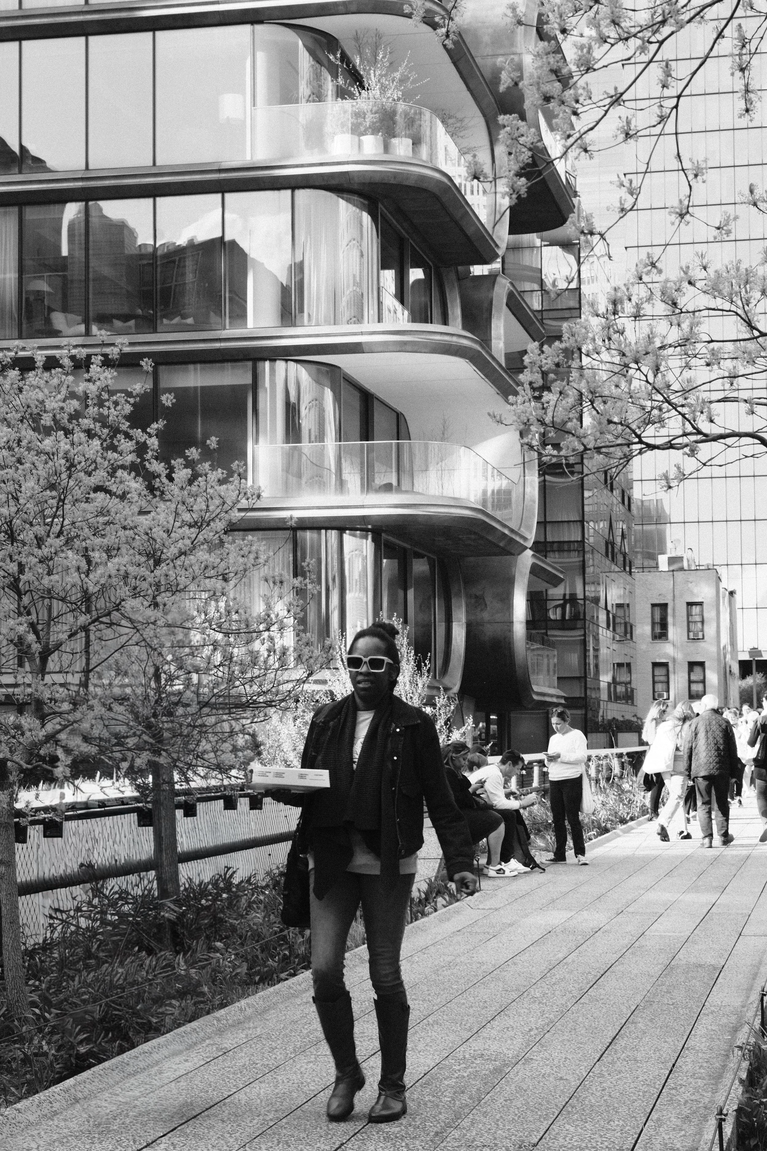 Black and white photo of a modern apartment building with glass balconies, people walking and sitting on benches, and a woman with sunglasses walking on a sidewalk carrying a pizza box.