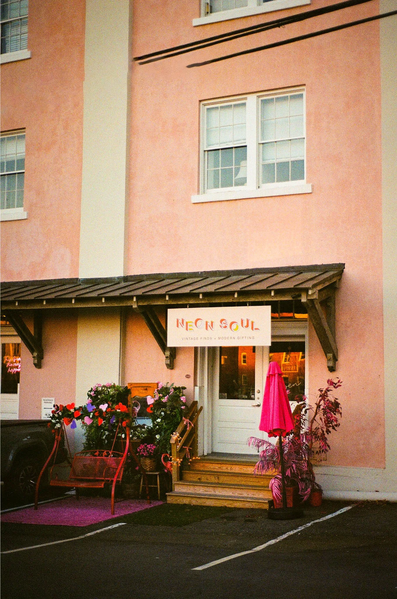The exterior of a pink building with a shop called Neon Soul. The shop has a sign, potted plants, flowers, and pink decor outside. There are parking spaces in front.