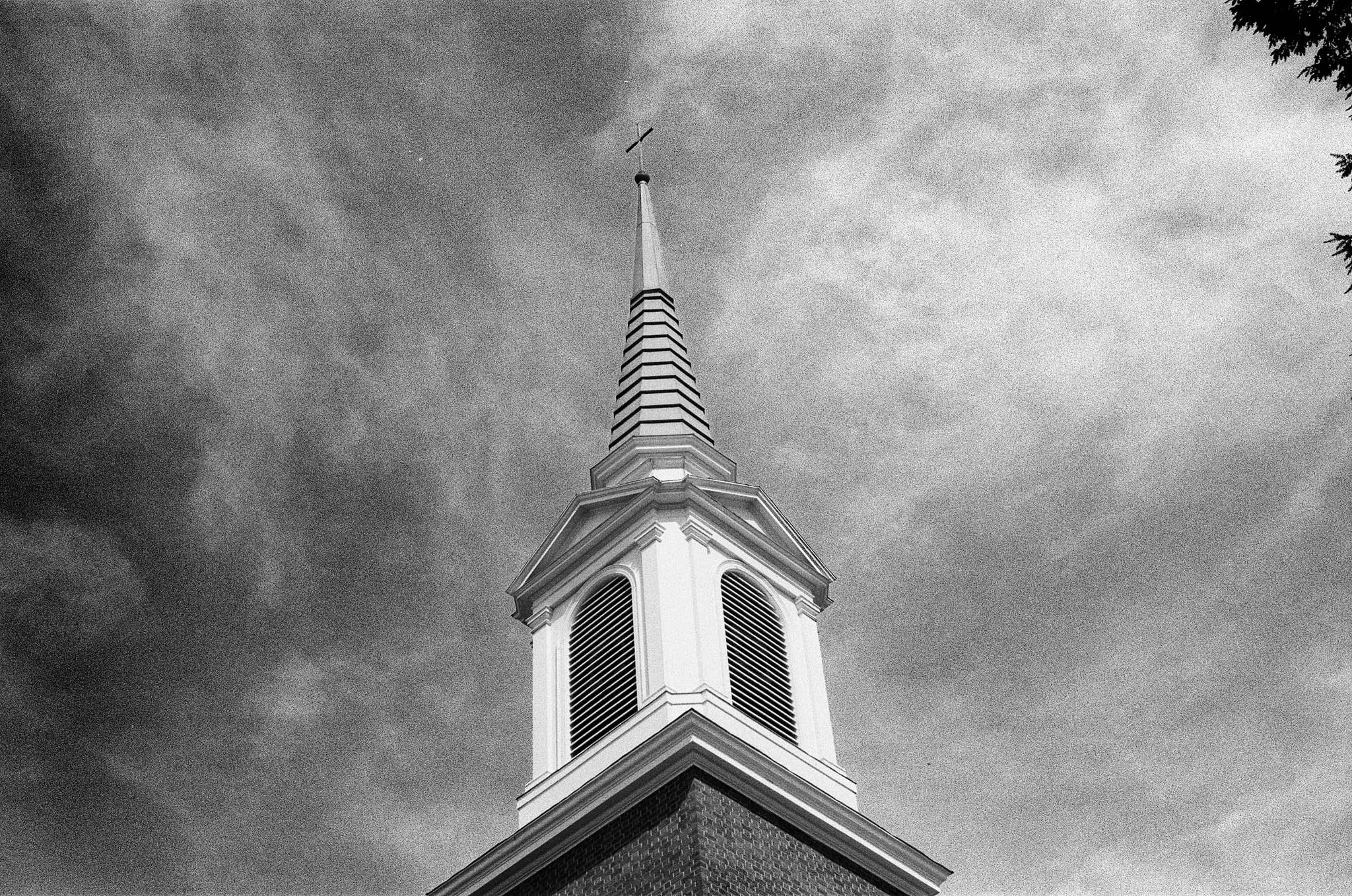 Black and white photo of a church steeple with a cross on top, set against a cloudy sky.
