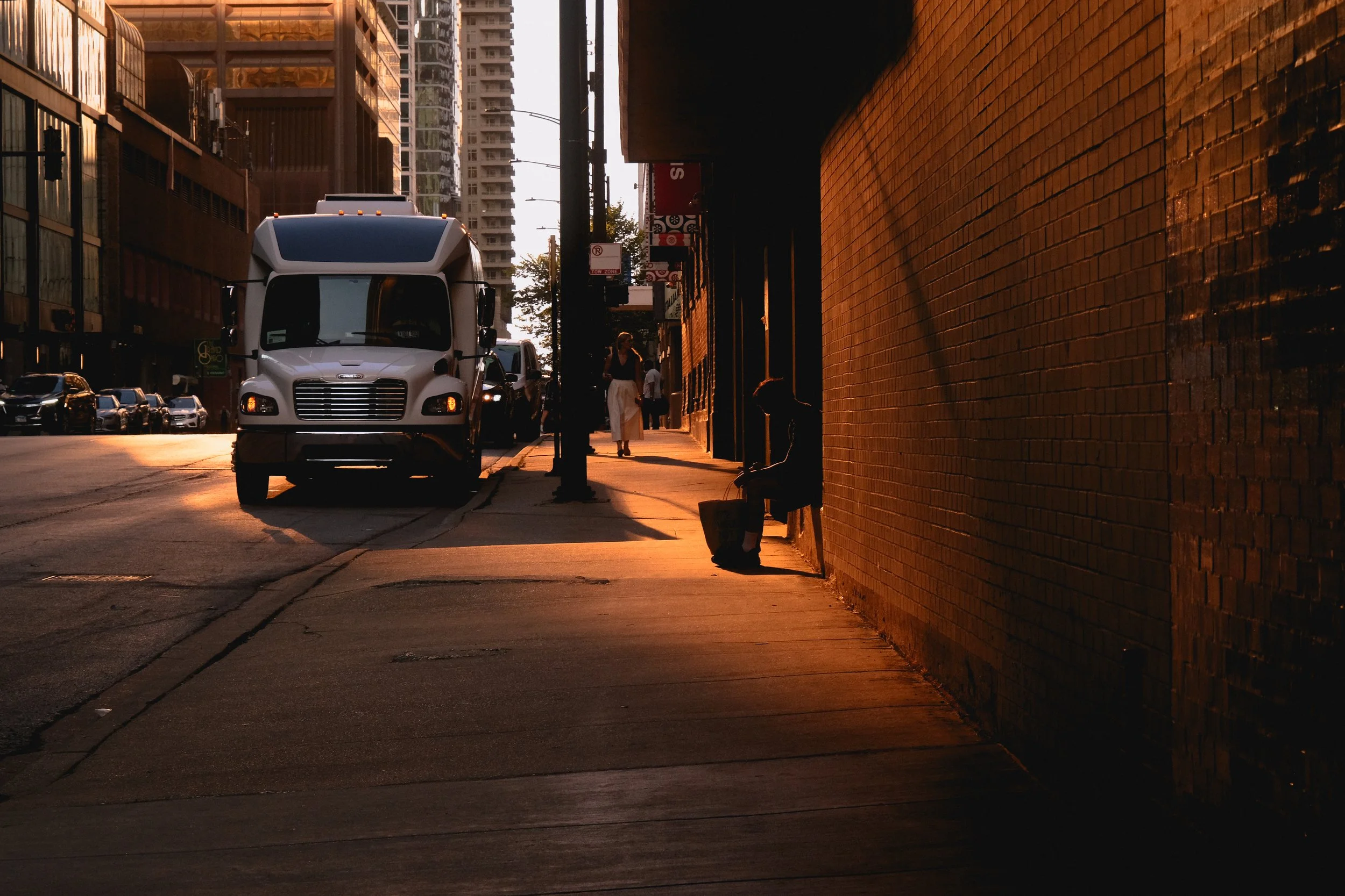 A city street scene during sunset with a white delivery truck, pedestrians walking, and a person sitting on a bench against a brick wall, illuminated by orange light.