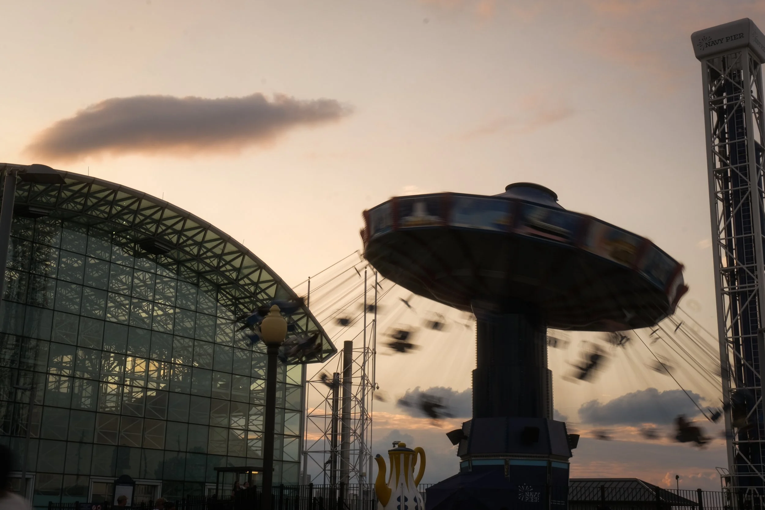An amusement park ride spinning at sunset with a modern glass building and a teapot sculpture in the foreground.