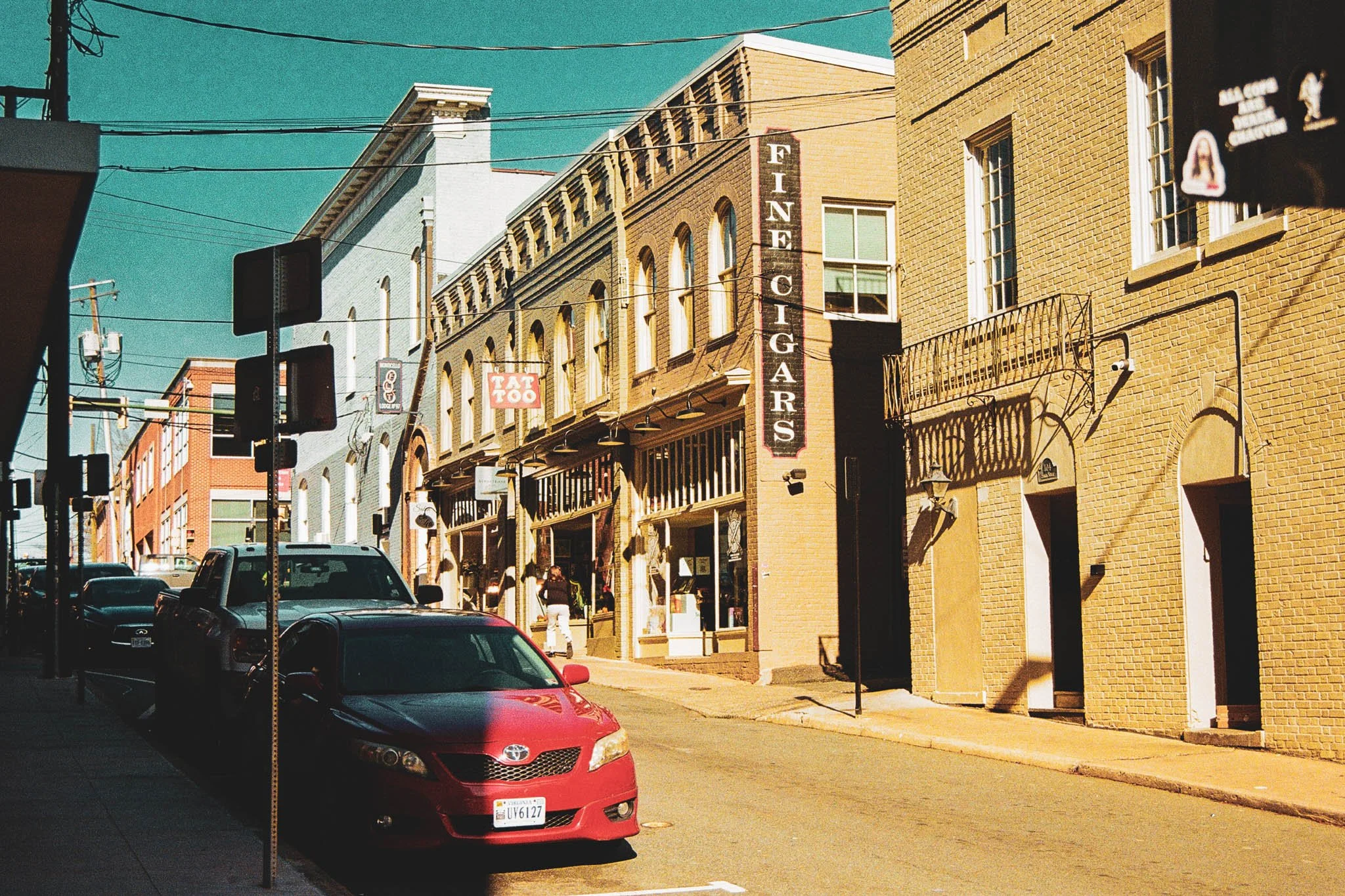 Street scene with parked cars, brick buildings, and signs for a tattoo shop and fine cigars under a clear sky.