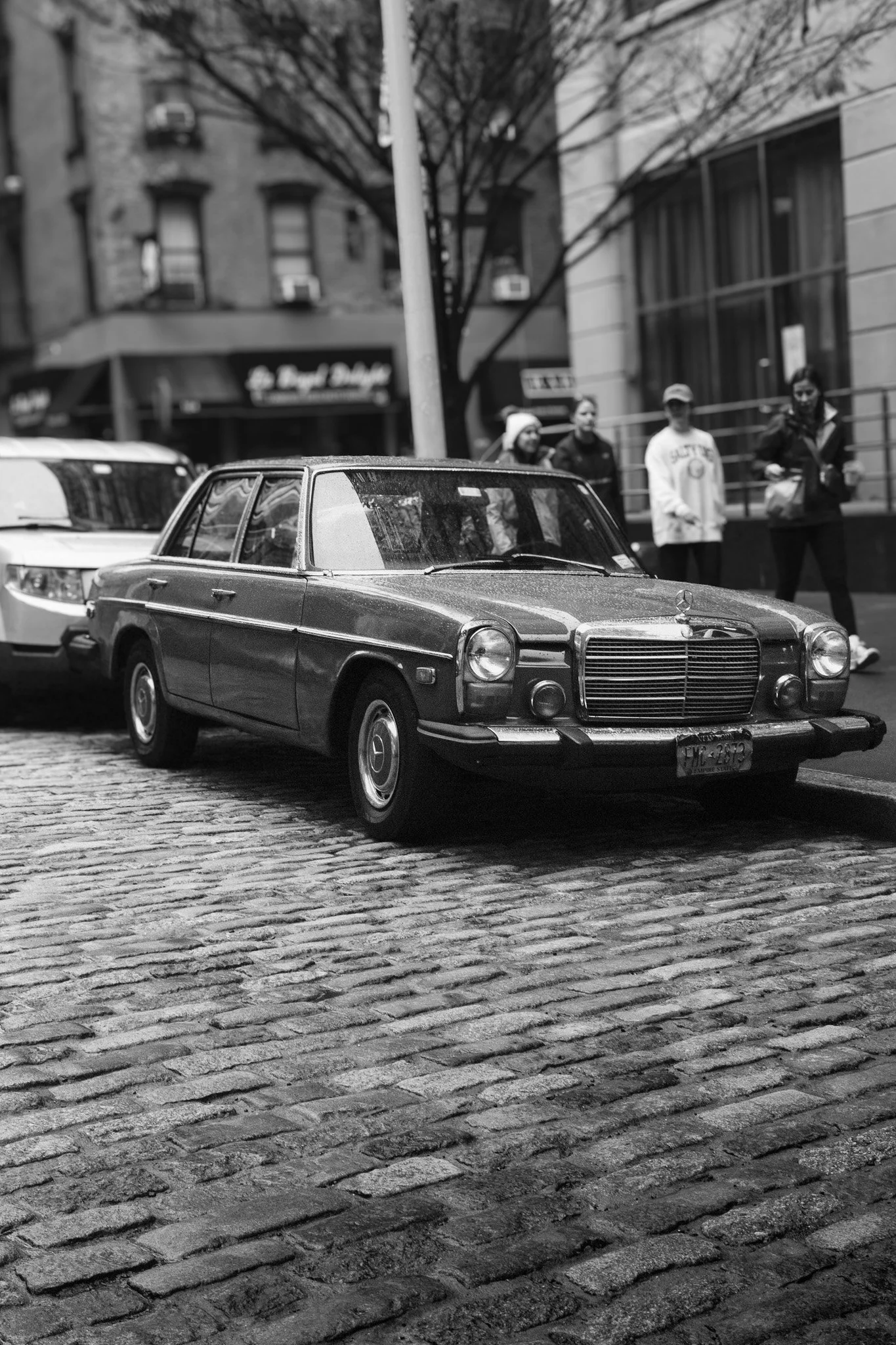 A black-and-white photo of a vintage Mercedes-Benz car parked on a cobblestone street with a group of people standing in the background.