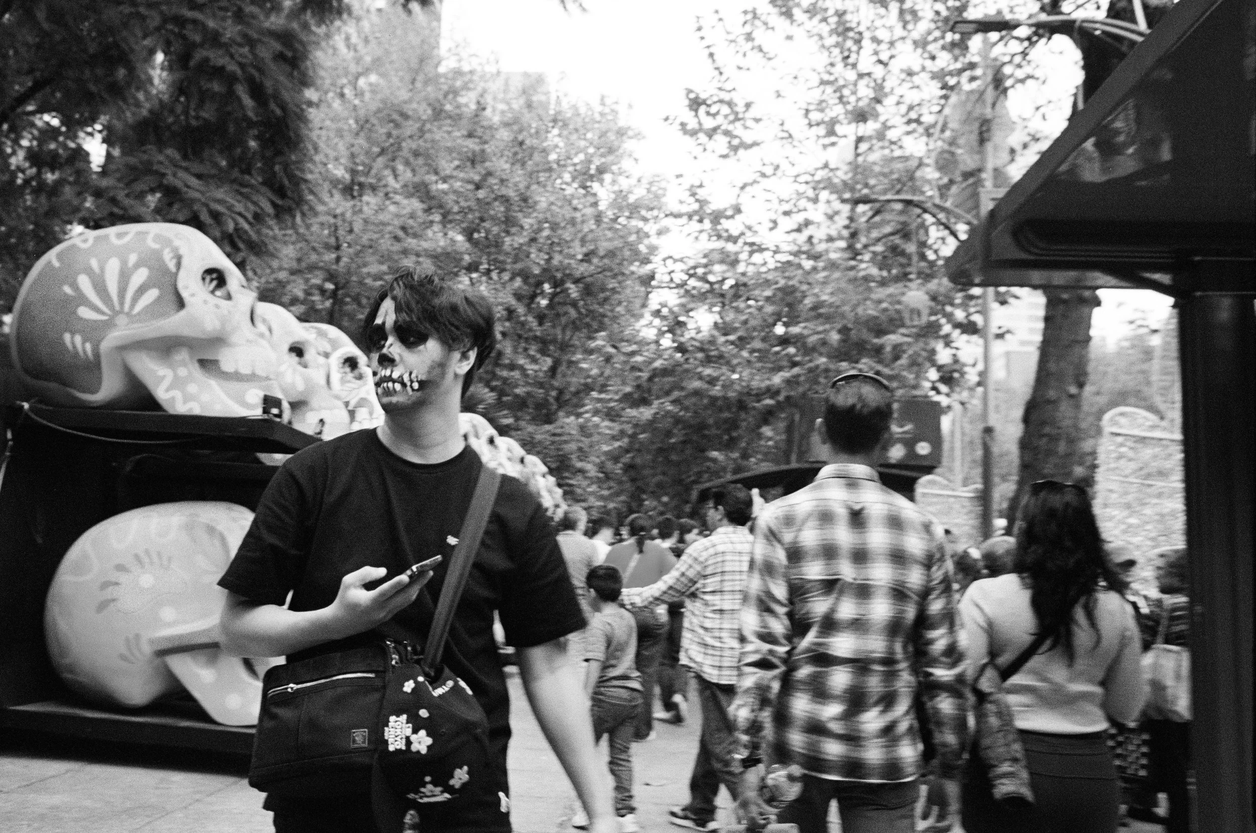 A person with face paint resembling a skeleton, standing in front of sugar skull decorations at an outdoor market or festival, surrounded by other people walking among trees.
