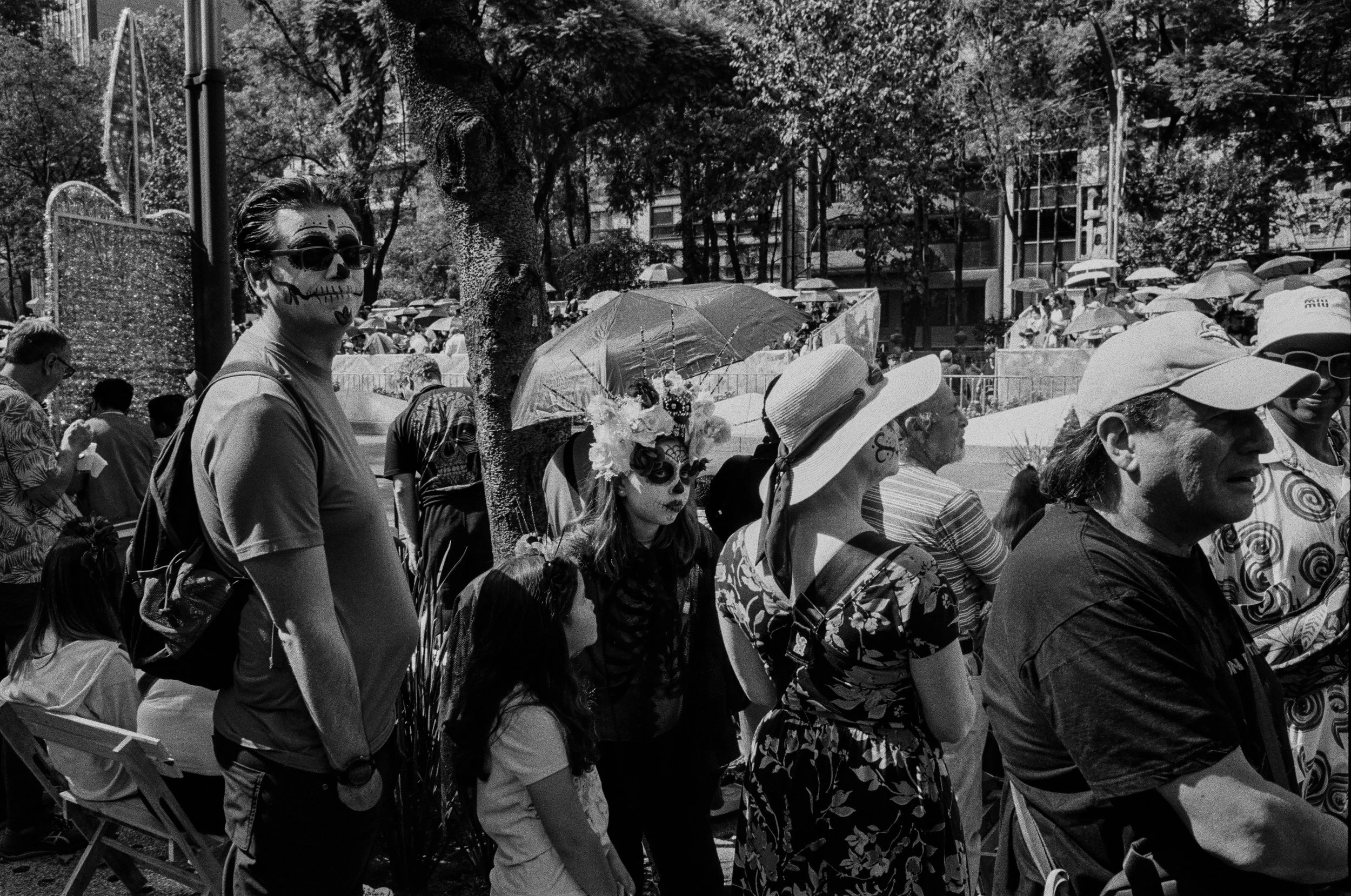 People gathered outside for a celebration or event, some with faces painted in sugar skull designs, wearing hats and costumes, some with umbrellas, in a park setting.
