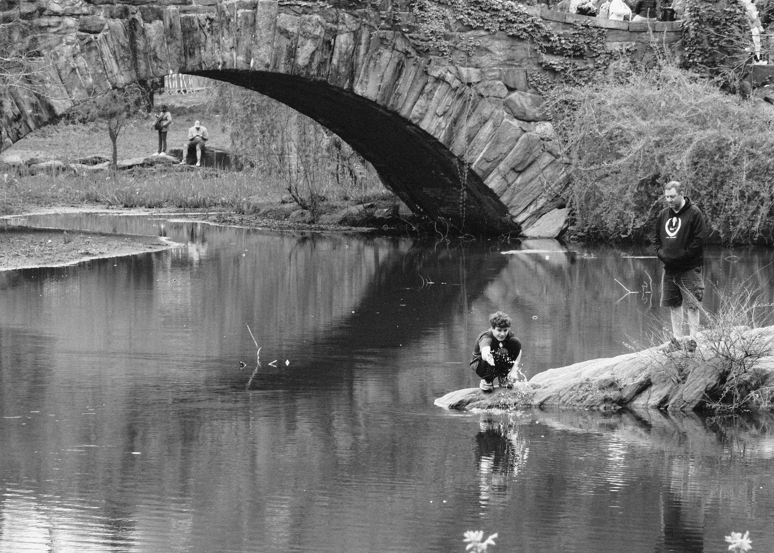 A black and white photo of a young boy squatting on a rock in a river, splashing water with his hand. An adult man stands nearby on the same rock, watching. In the background, there is an arched stone bridge and two people sitting on a bench undernea