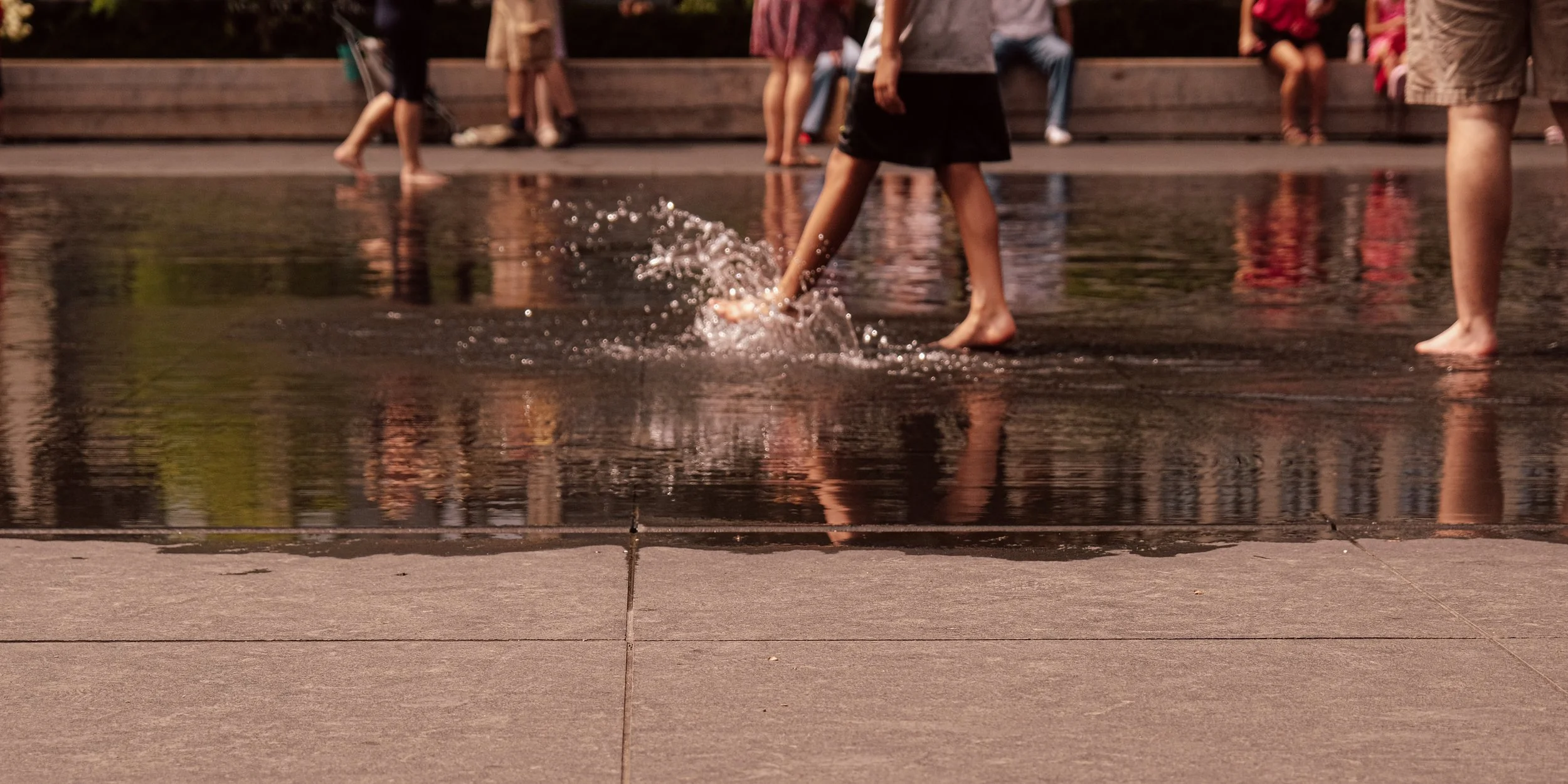 Children playing in a shallow water fountain with ripples and splashes, surrounded by a concrete surface and people sitting on a wooden bench in the background.