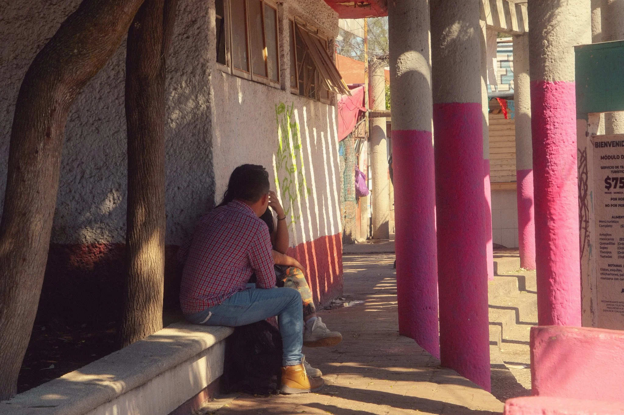 A young man and a young woman sit on a concrete bench under a tree on a city sidewalk, with colorful pink and blue painted columns nearby. They appear to be talking or resting.