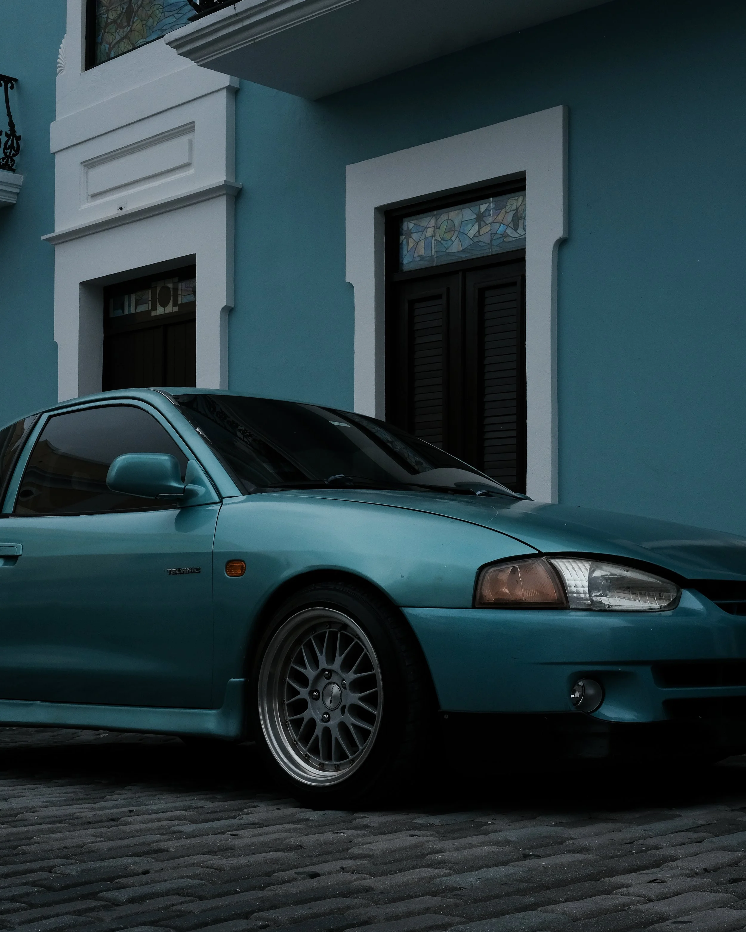 A teal-colored Honda Prelude parked in front of a blue house with white trim, cobblestone driveway, and stained glass window above the door.