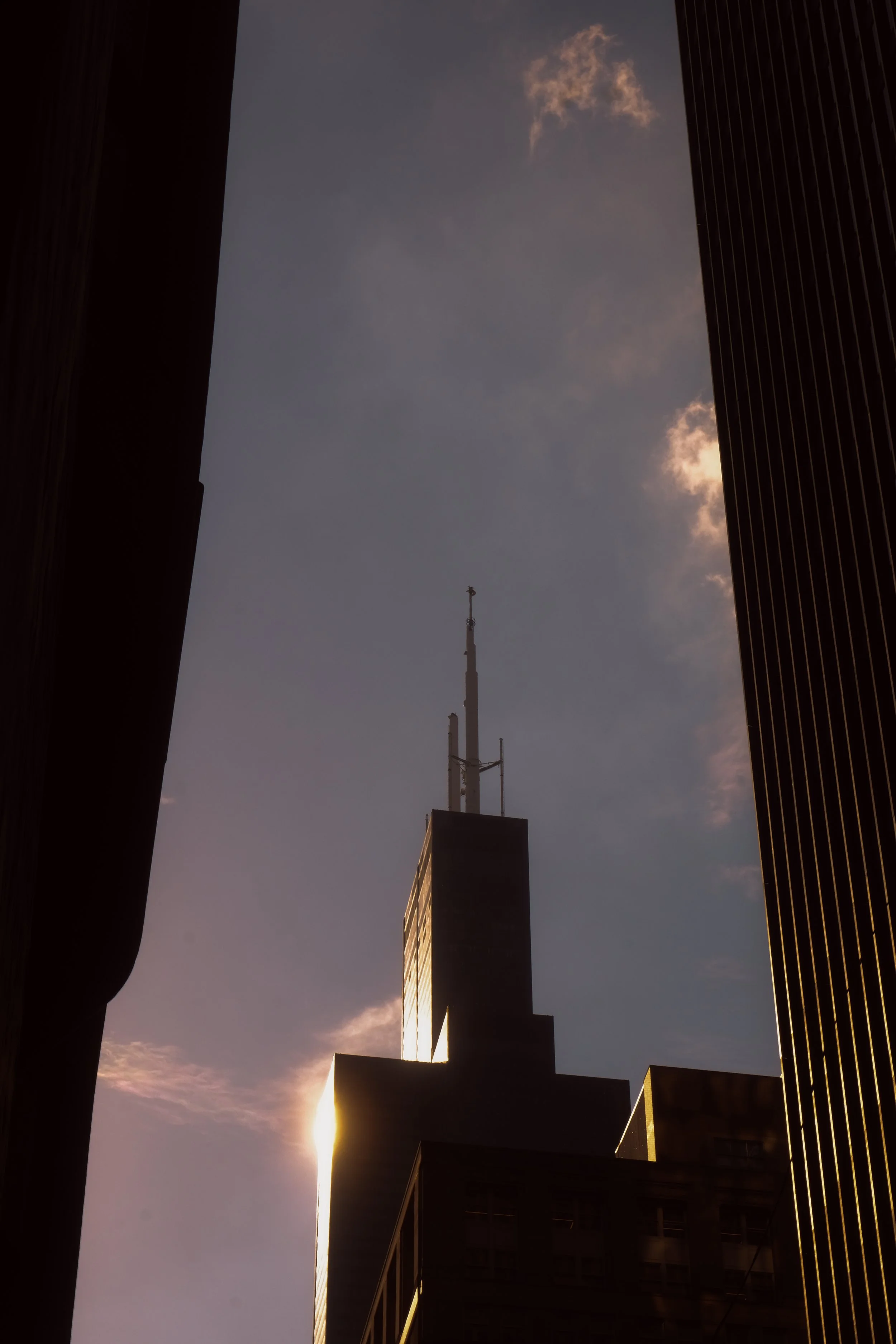 The photo shows the skyline of a city with a tall building in the center, featuring antenna towers on top. The building is partly illuminated by the setting or rising sun, creating a silhouette effect against the sky. There are dark silhouettes of ot
