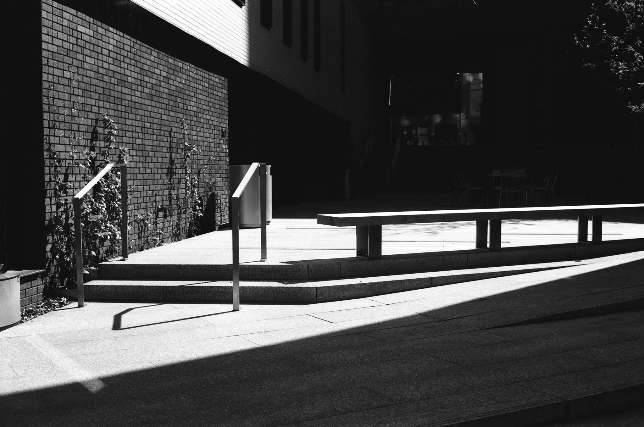 Empty outdoor area with stairs, railing, brick wall, benches, and shaded seating area in black and white.