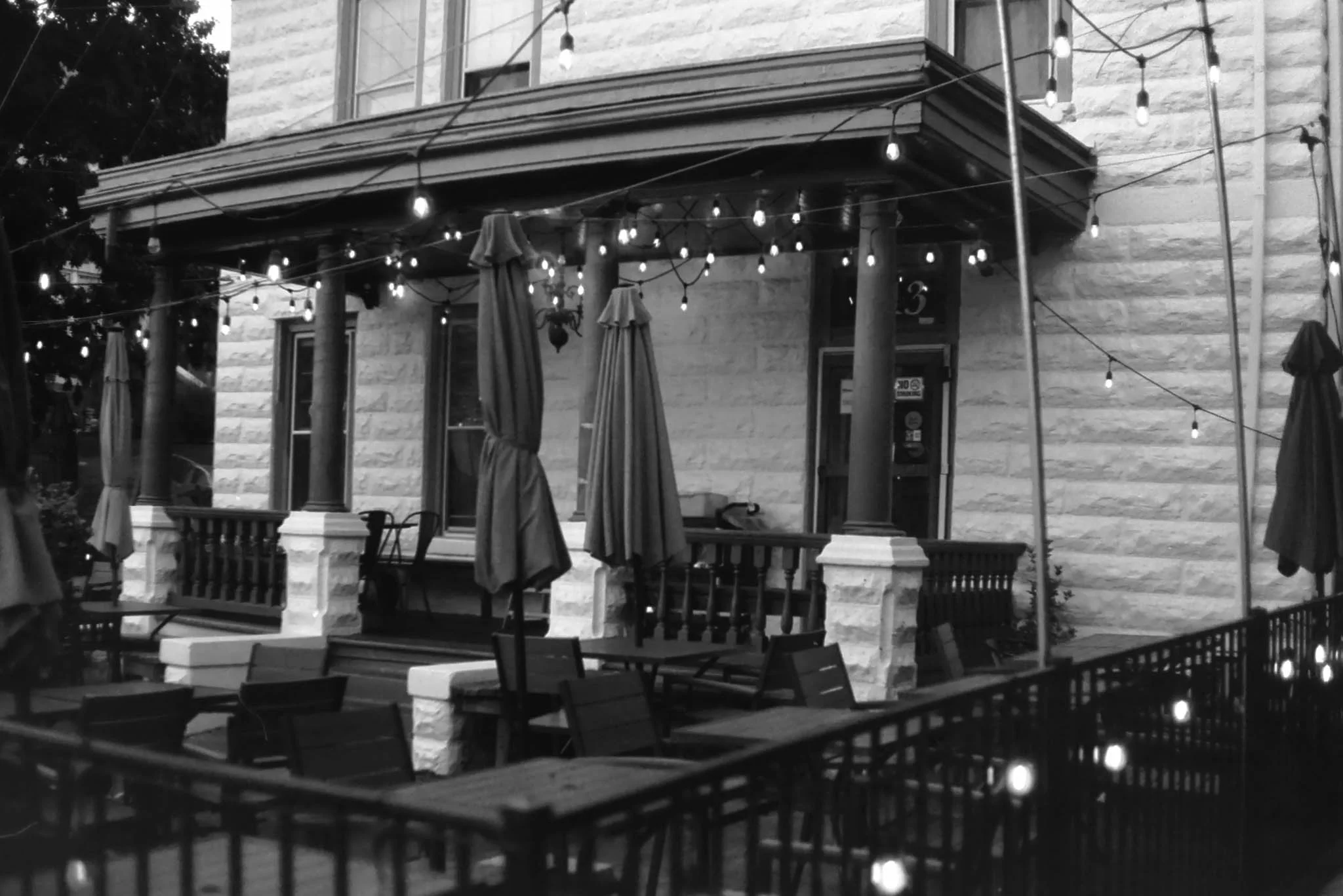 Black and white photo of an outdoor patio with tables, chairs, and umbrellas, decorated with string lights hanging overhead. A building with a stone facade and windows is in the background.