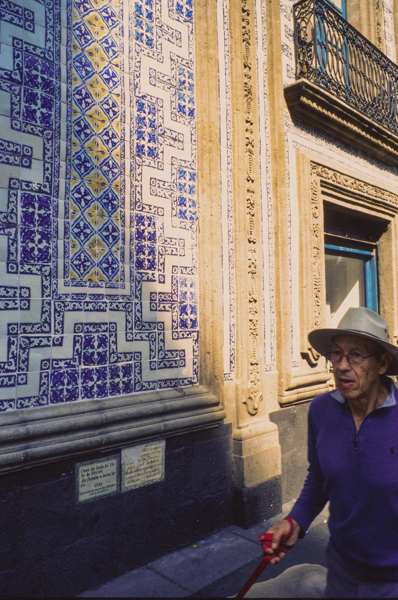 An elderly man wearing a light-colored hat, glasses, and a purple sweater walking past a building with intricate blue, white, and yellow tile work and decorative stone detailing.
