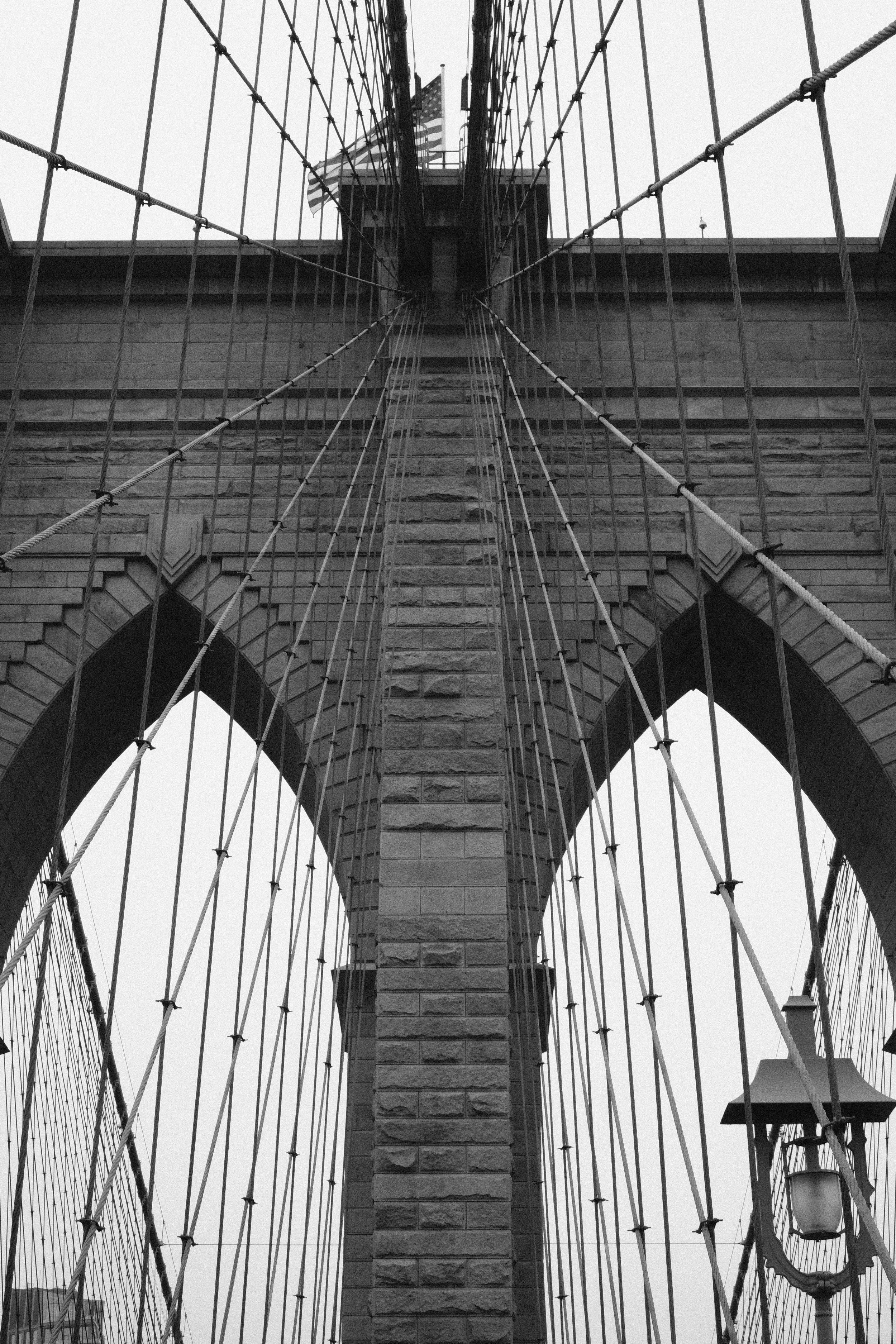 Upward view of the Brooklyn Bridge in New York City, showing stone arches and suspension cables with American flag in the background.