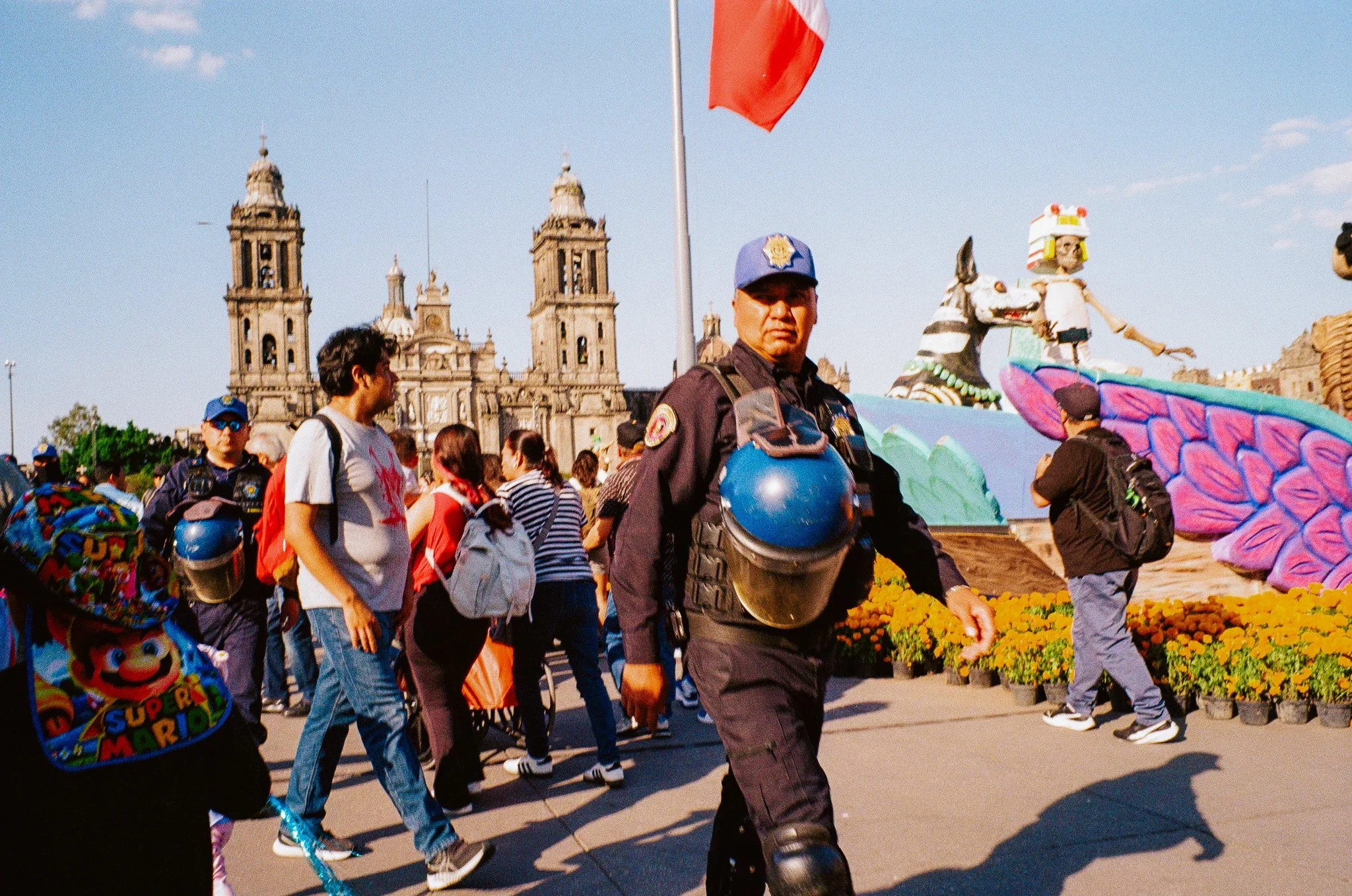 Police officers and pedestrians in a busy city square with large cathedral in the background and colorful parade floats.