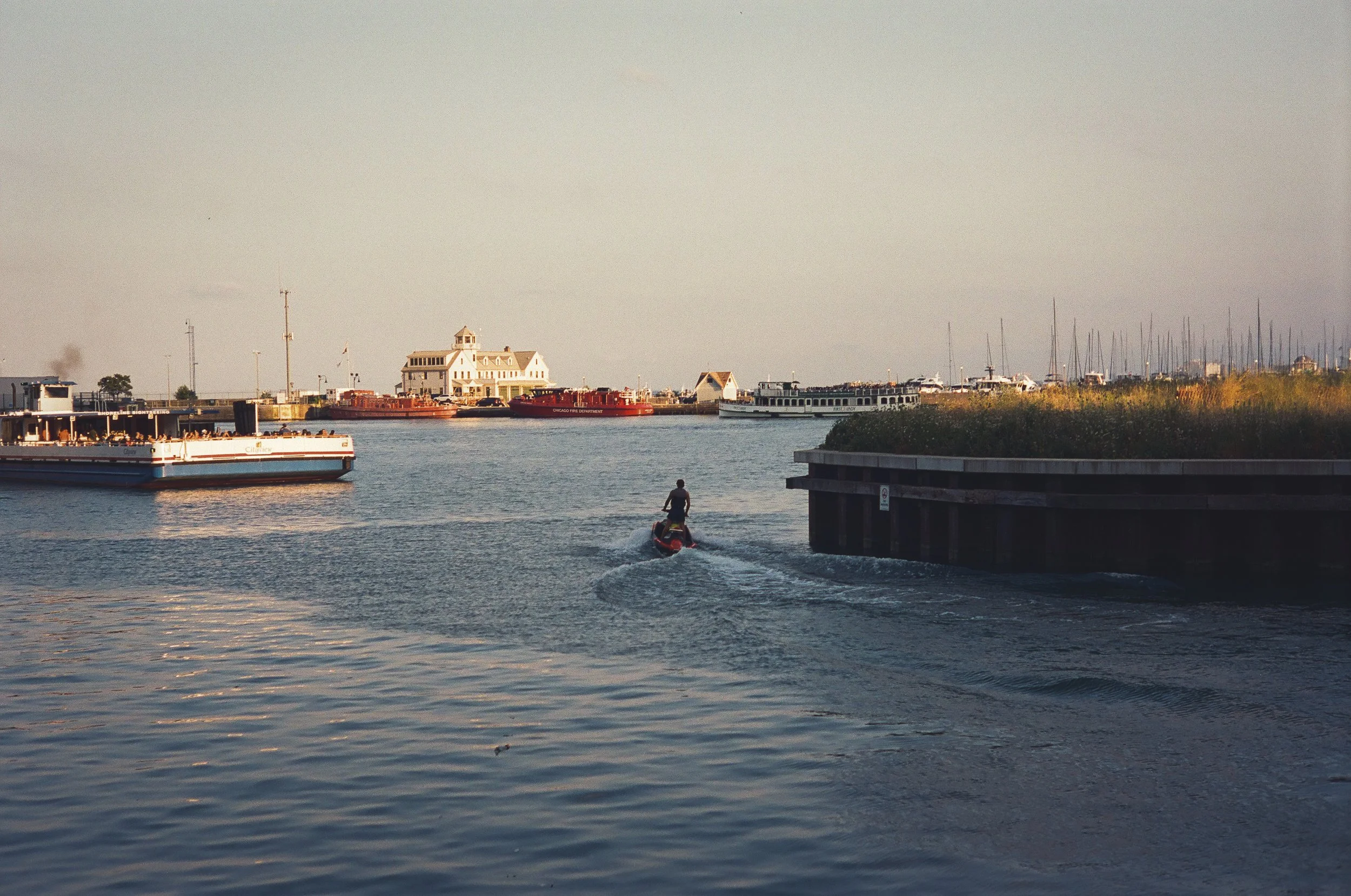 A person riding a jet ski on a calm harbor with boats docked on the sides and a large house on the far shoreline in the background, under a clear sky.