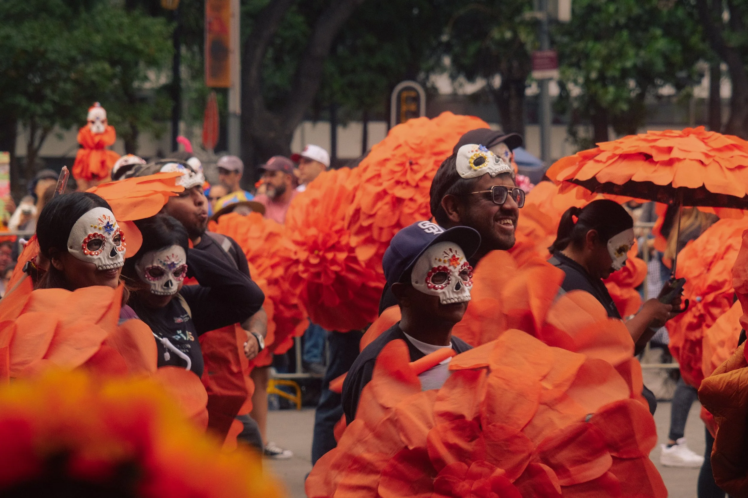 People dressed in colorful costumes with skull face paint, participating in a street celebration, with orange decorations and tissue paper flowers, possibly for Day of the Dead.