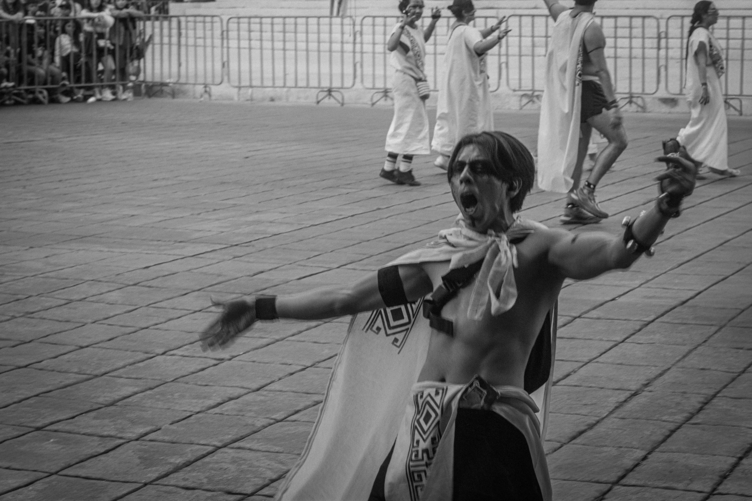 A shirtless man with long hair, face paint, and a cape, shouting passionately and extending his arm during a parade or celebration.