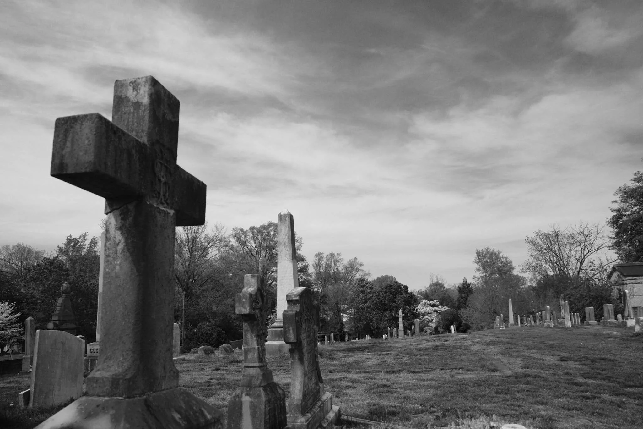 Black and white photo of a cemetery with various tombstones and crosses, trees in the background, and a cloudy sky.