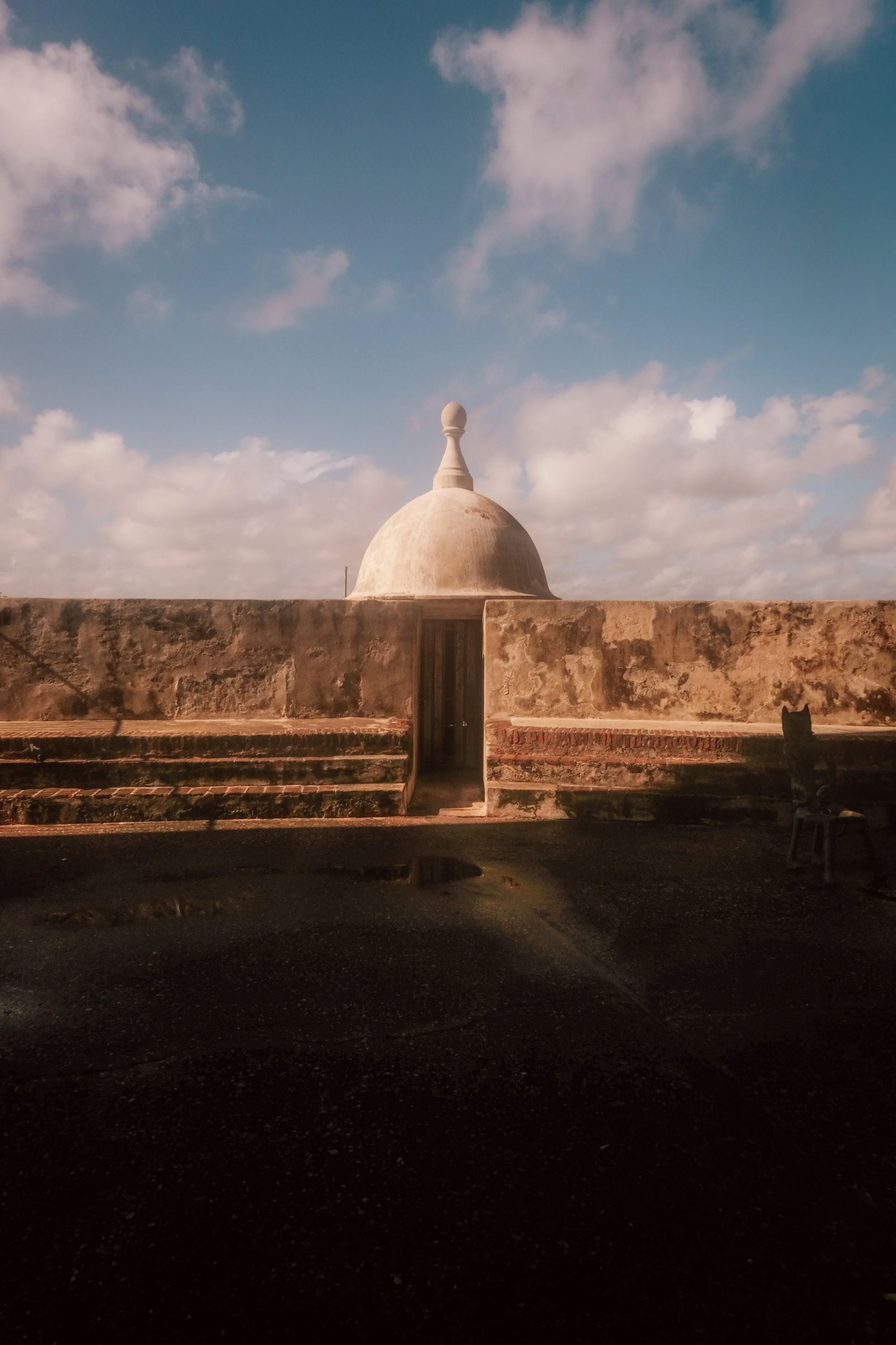 A historical structure with a domed roof, possibly a small fortress or temple, with a stairway leading up to it, set against a partly cloudy sky.