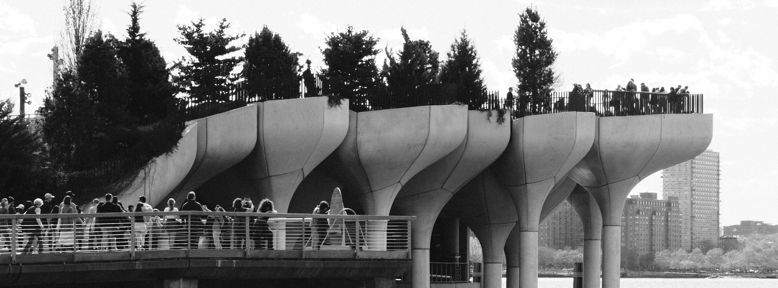 People walking and standing on a bridge with an elevated park structure with trees on top, city buildings in the background