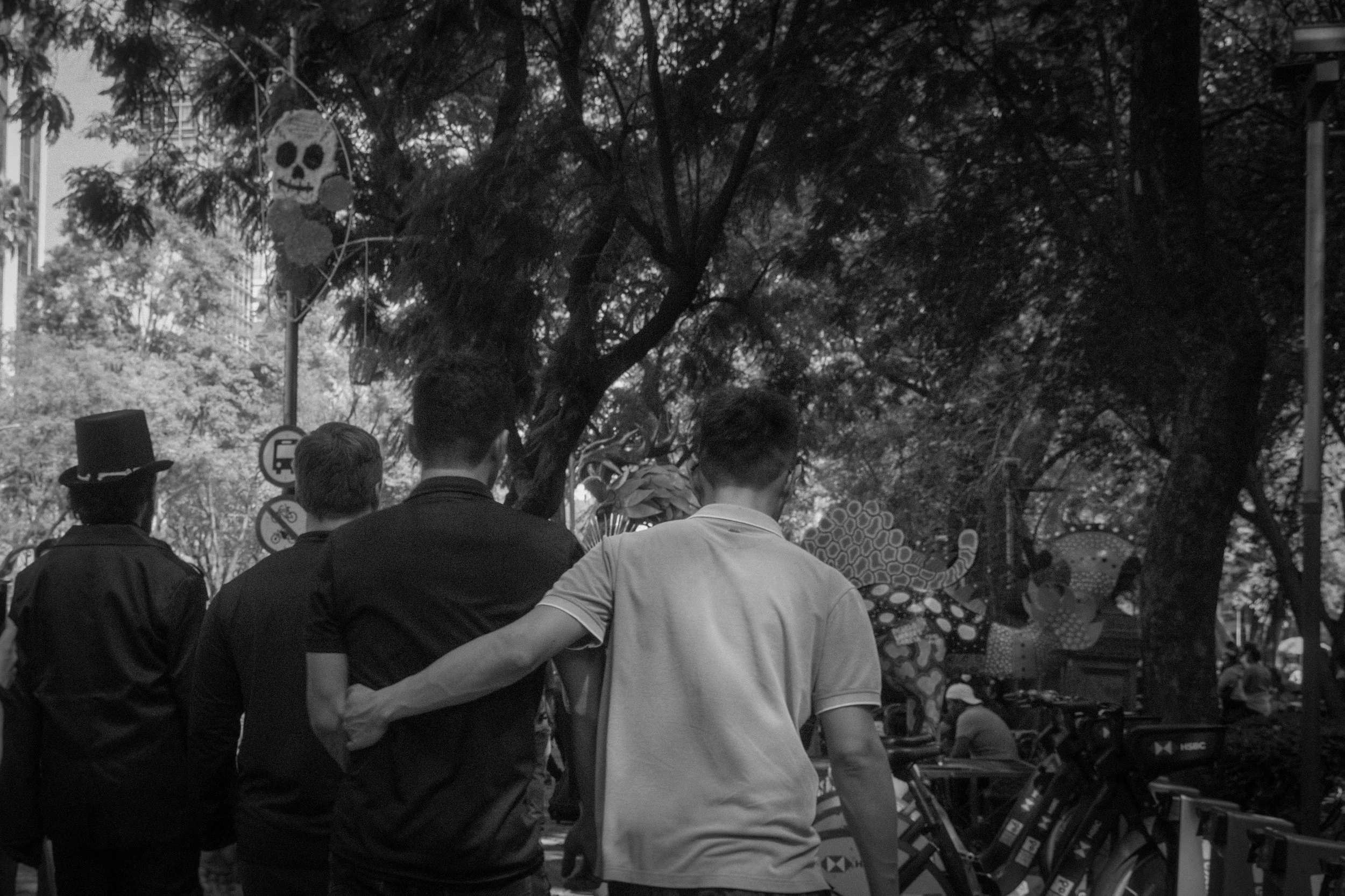 A black-and-white photo showing three men walking in a park, one of whom is holding onto another's shoulder. There are bicycles parked to the right and a decoration of a skull and flowers hanging from a tree in the background.
