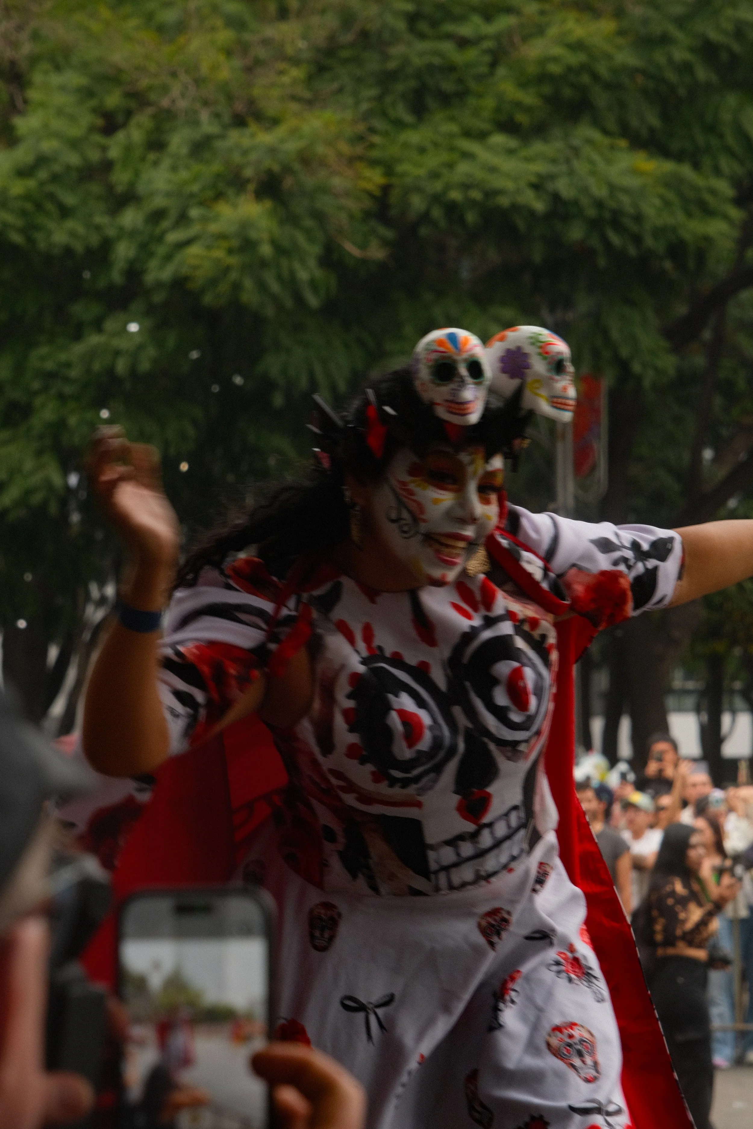 A woman dressed in traditional Mexican Day of the Dead costume with face paint, wearing a dress with sugar skull designs, dancing among a crowd with trees in the background.