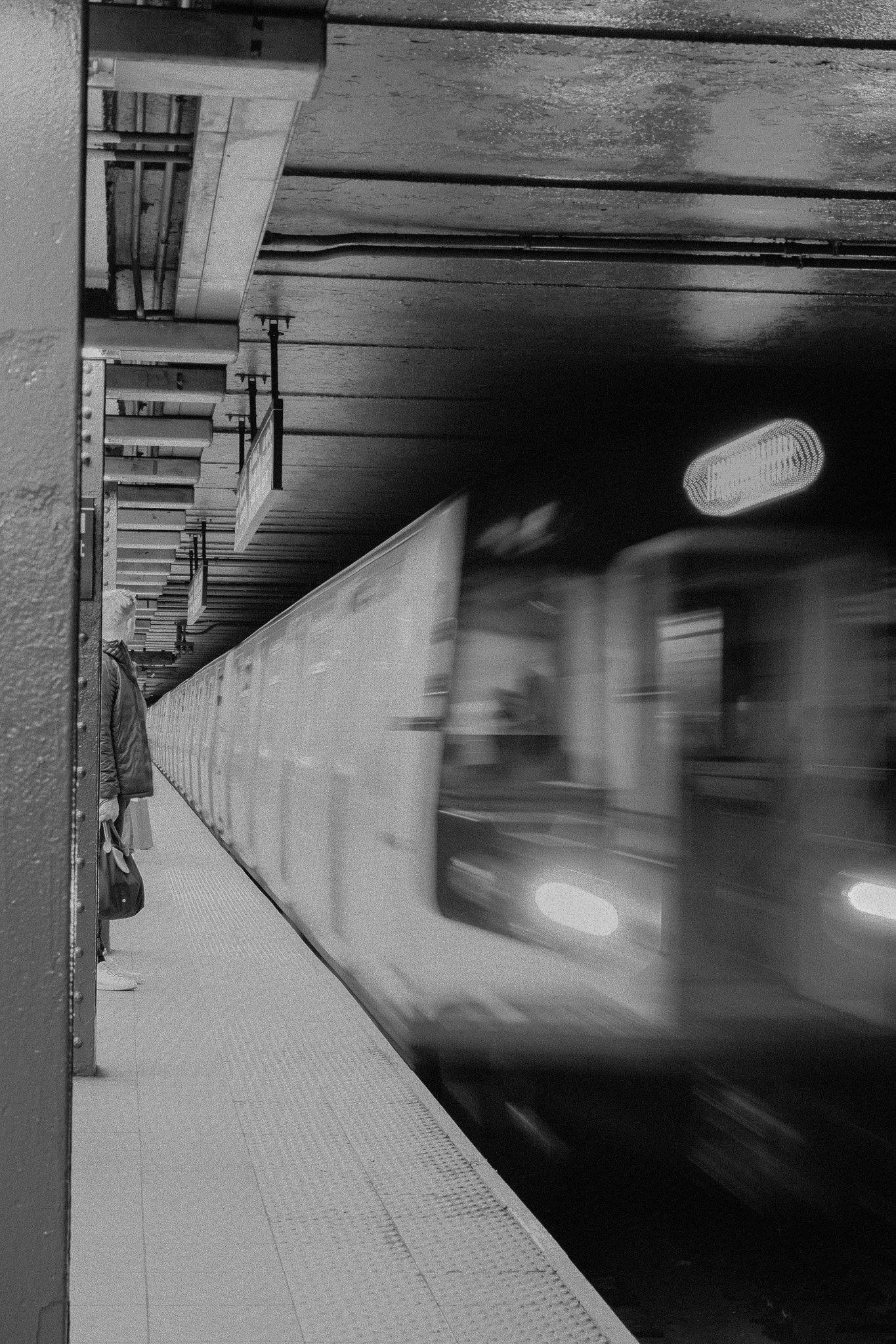 A subway train arriving at a station platform with a person standing and waiting, in black and white.