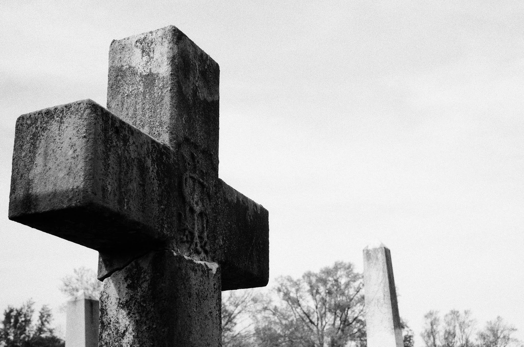 Black and white photograph of a weathered stone cross monument with trees and other crosses in the background.