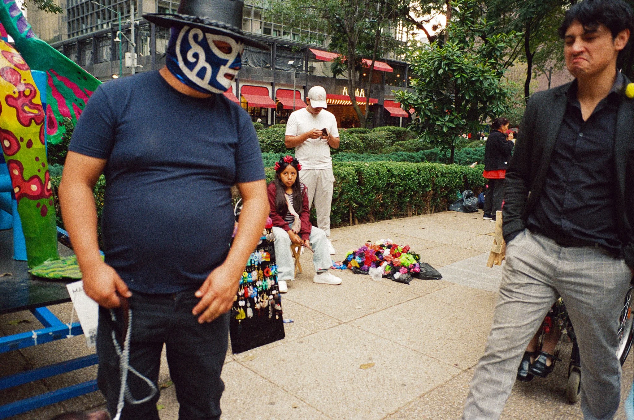 People at an outdoor market with vendors and flowers, including a woman wearing a traditional Mexican mask, a young girl with flowers in her hair, a man using his phone, and a man with a pained expression wearing a black blazer and plaid pants.