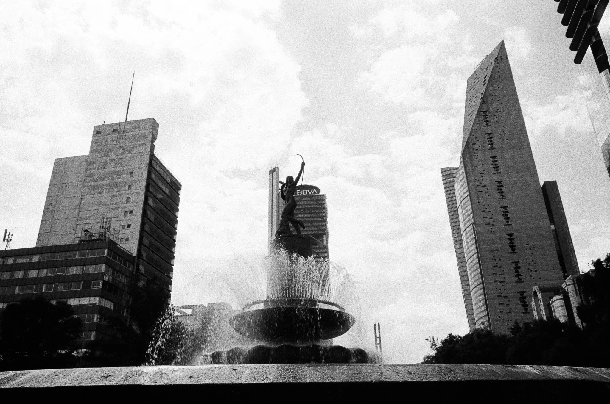 Black and white photo of a city square with tall modern buildings surrounding a fountain with a statue of a woman holding a bow and arrow.