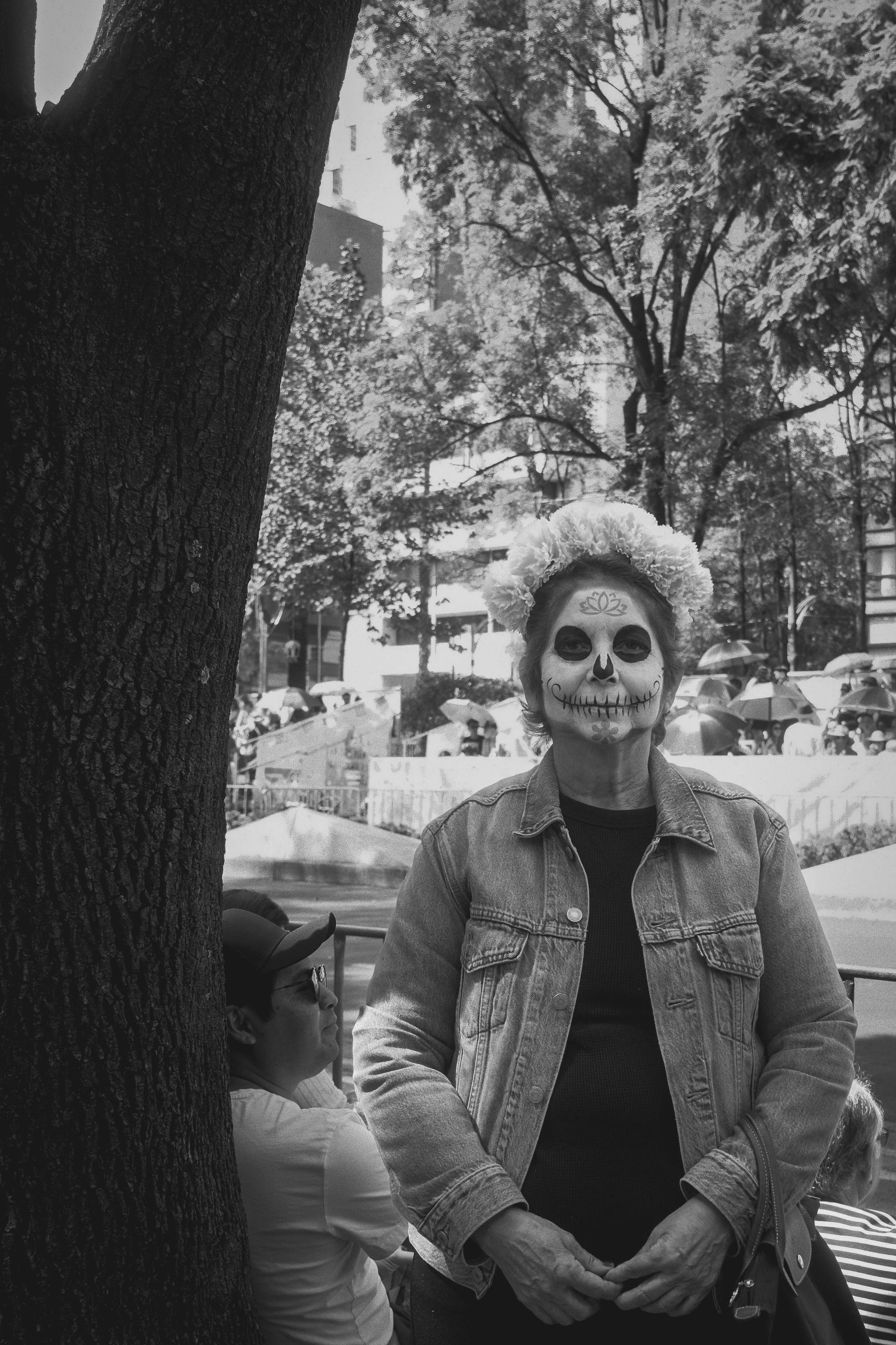 Person with sugar skull face paint or makeup, wearing a floral headpiece and denim jacket, standing outdoors among trees and people with umbrellas.