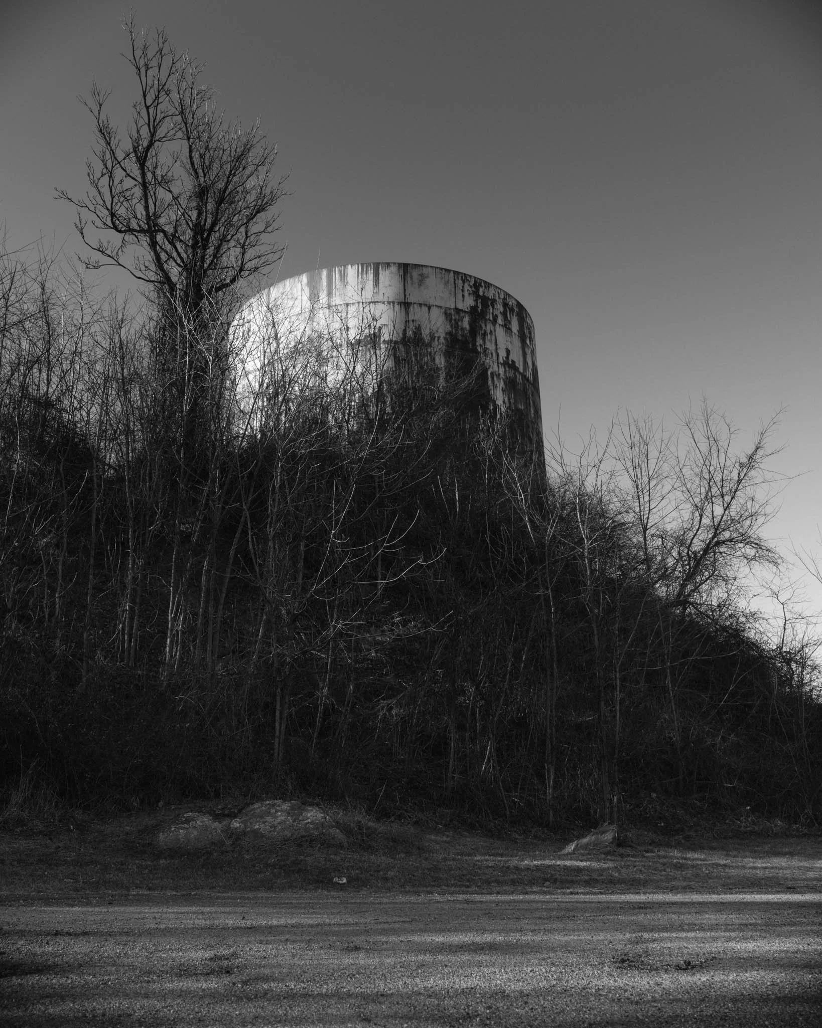 An old, rusted, cylindrical water tower surrounded by leafless trees on a hill, with a gravel road in the foreground, in black and white.