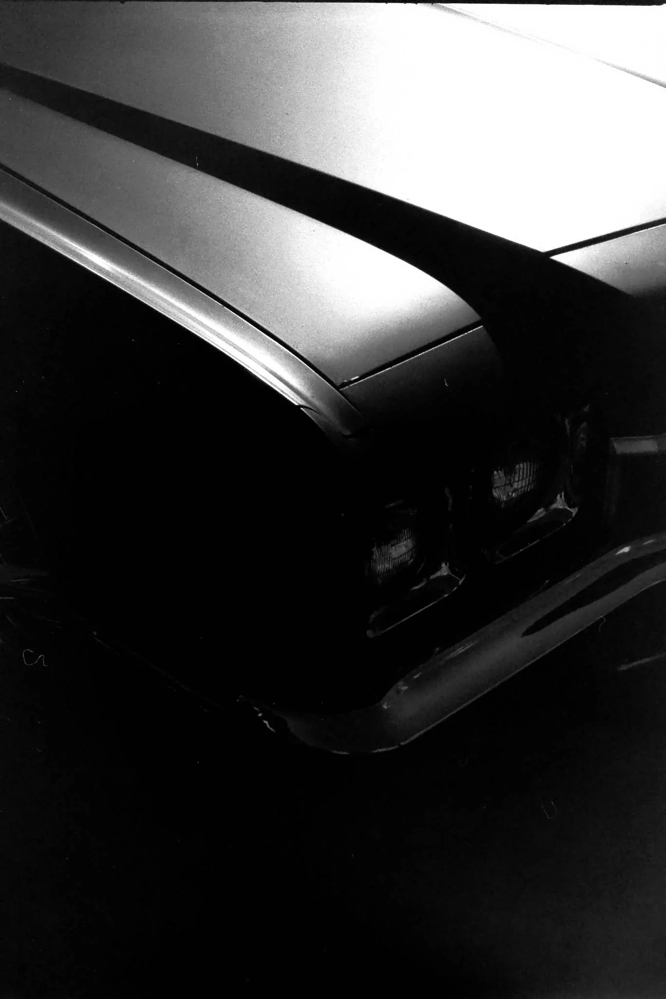 Close-up of the front of a classic car, showing the headlight, hood, and part of the bumper in black and white.