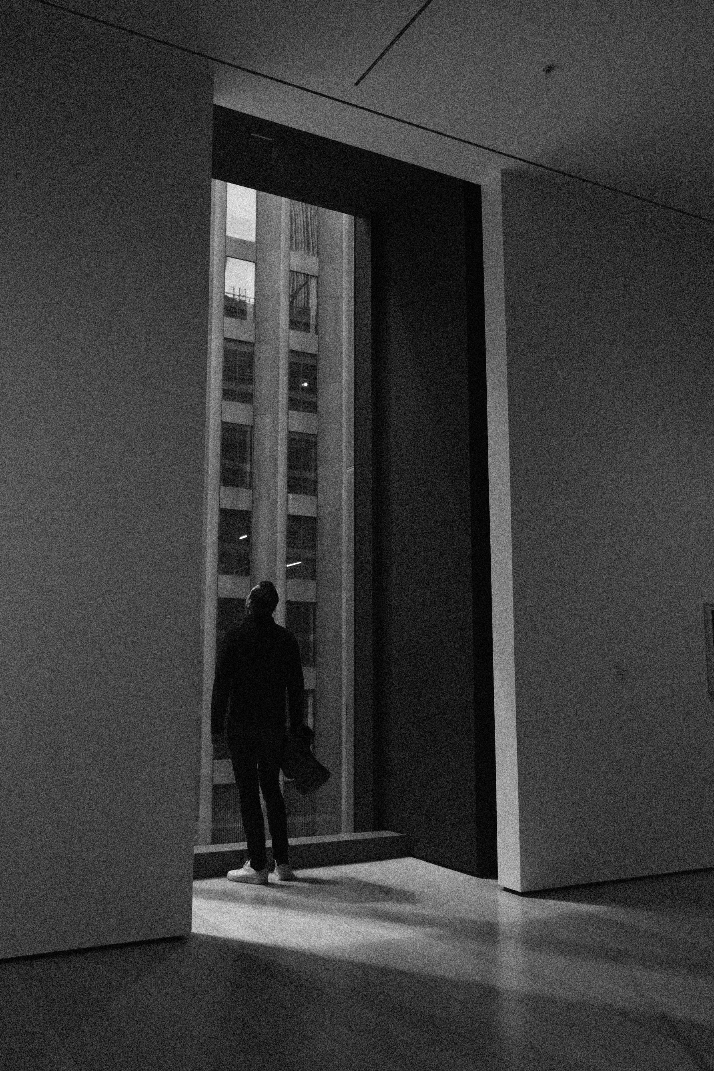 A person standing inside a modern building, looking out of a tall window onto an exterior high-rise building.