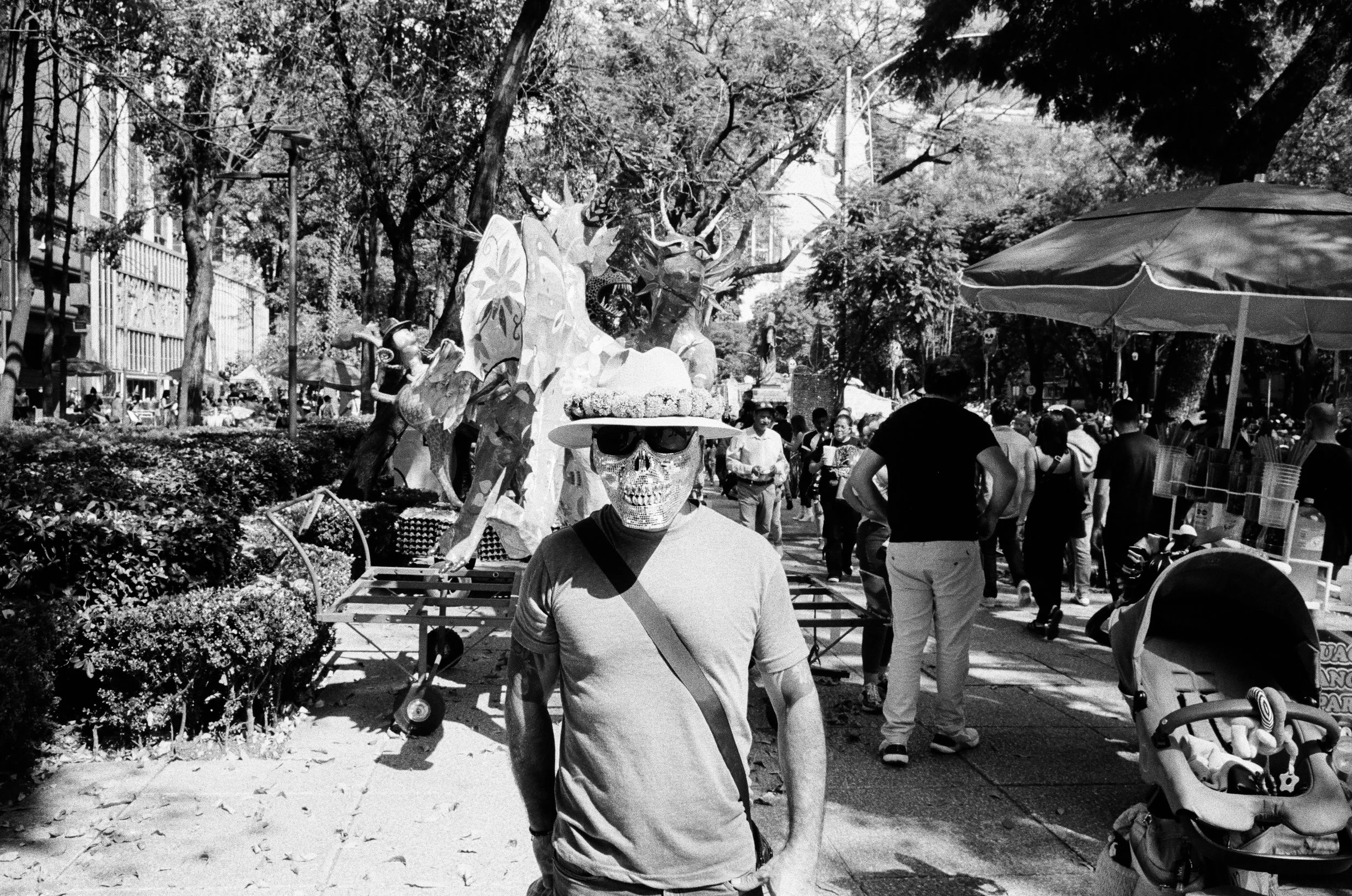 Person wearing a skull mask, sunglasses, and a floral hat, standing in a busy outdoor market.