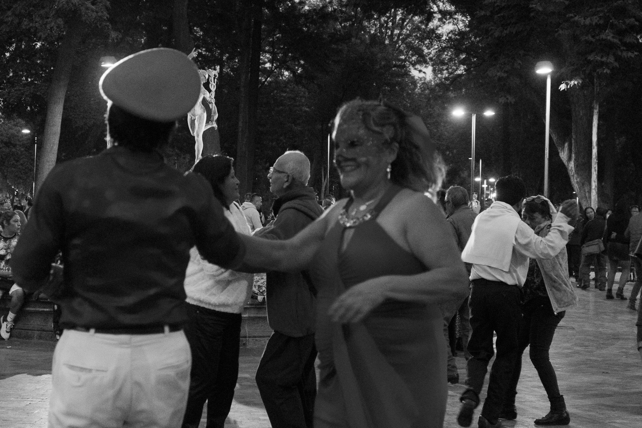 People dancing outdoors at night, with trees and streetlights in the background, in a lively social gathering.