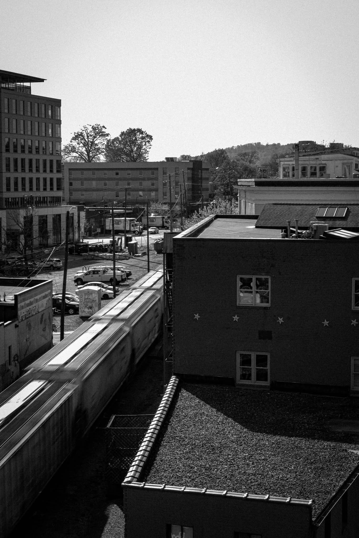 Black and white photo of a cityscape with buildings, parked cars, utility poles, and a moving train.
