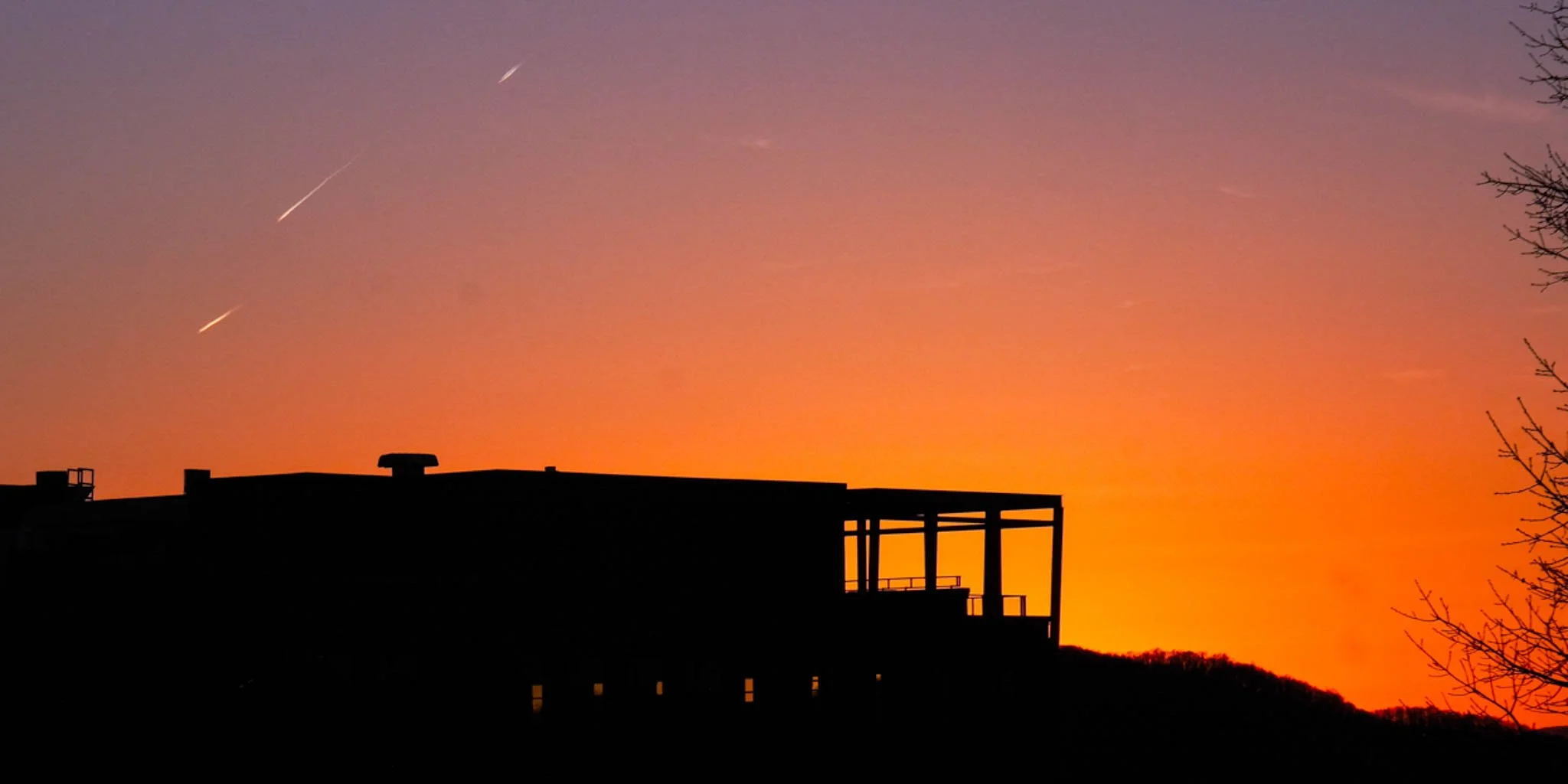 Silhouette of a building with an open balcony against a colorful sunset sky with streaks of light and a few clouds.