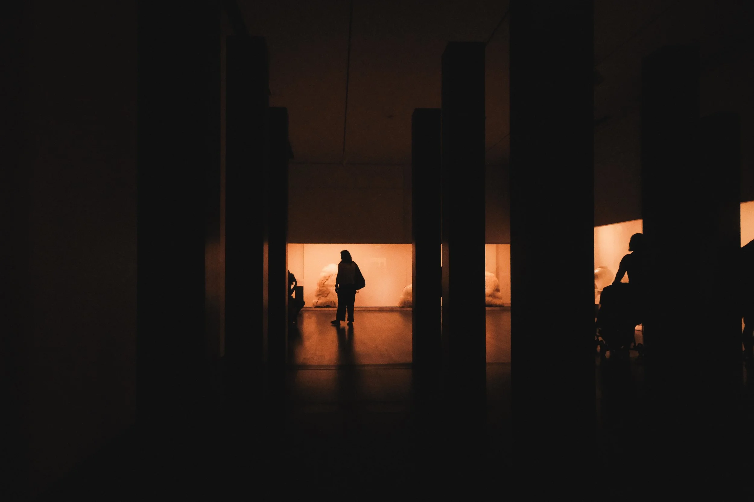 Silhouettes of people sitting and standing in a dimly lit gallery with bright artwork illuminated in the background.