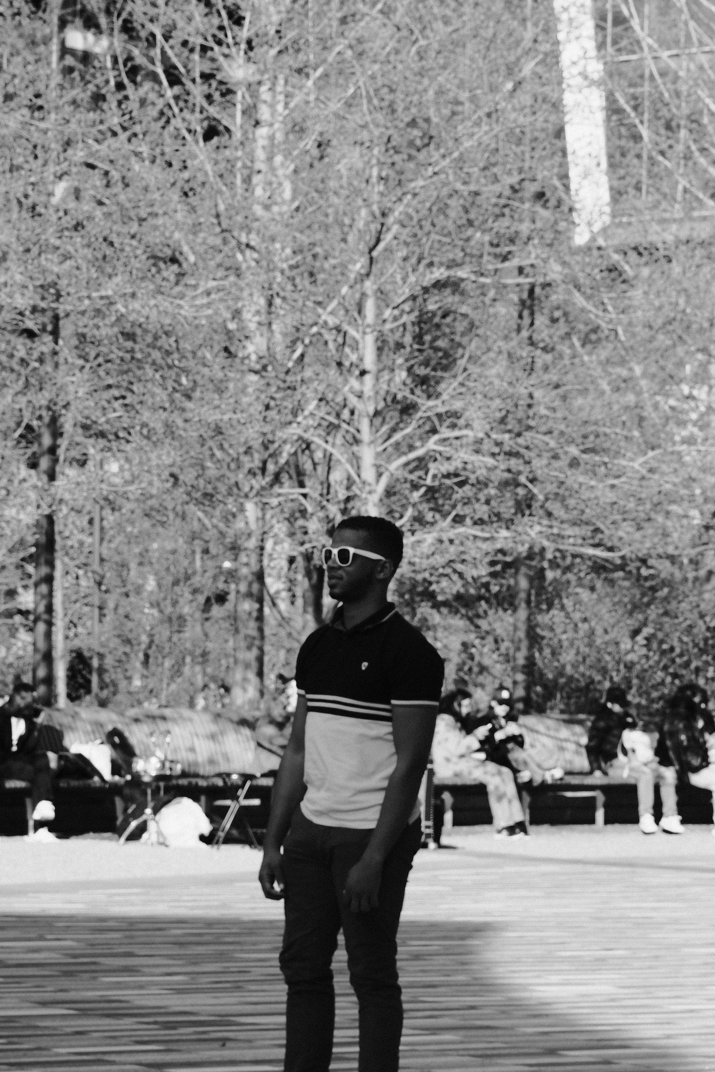 A young man wearing sunglasses and a black and white shirt standing outdoors in front of leafless trees, with people sitting on benches in the background.