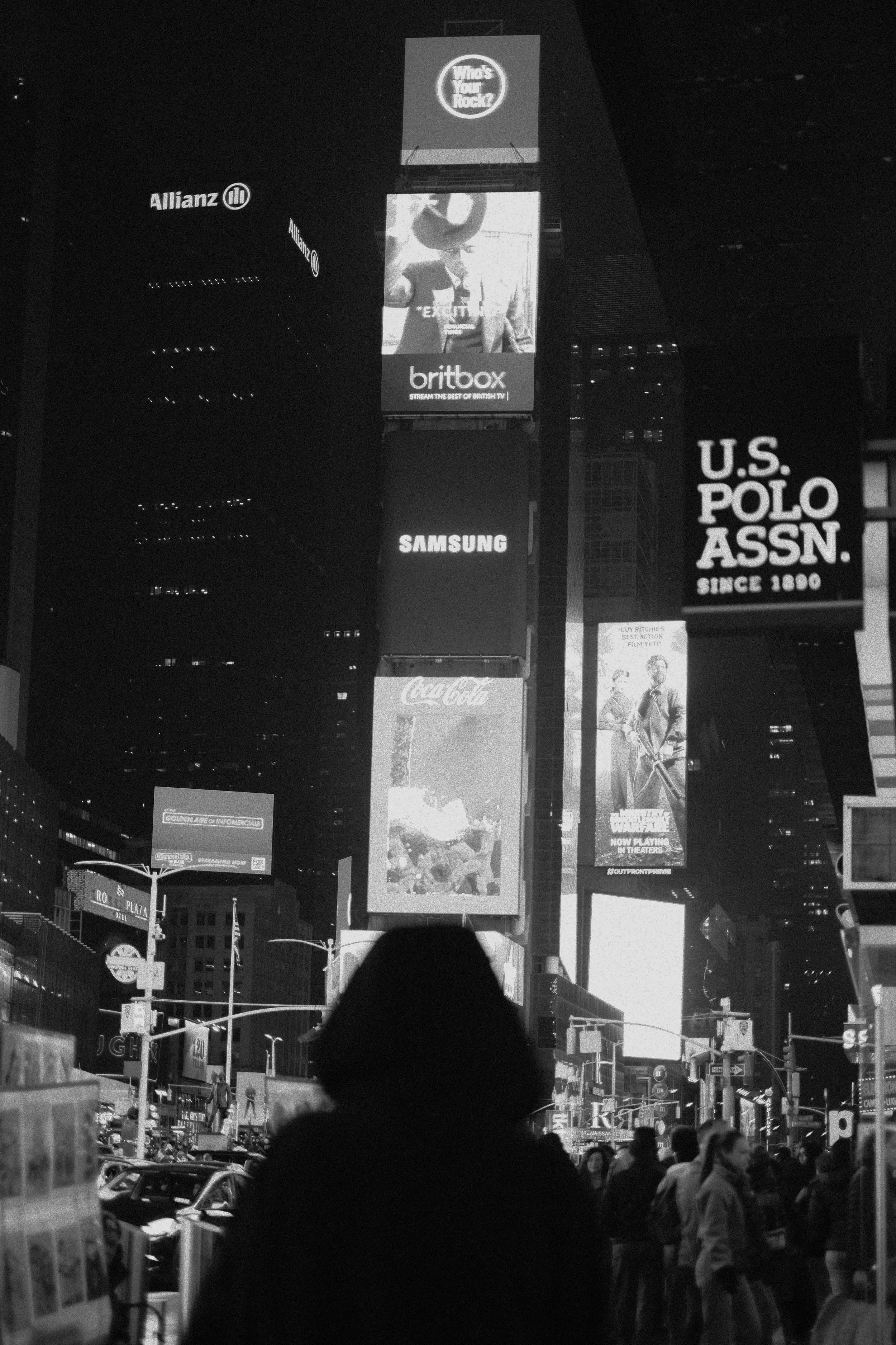 Nighttime scene in Times Square, New York City with illuminated billboards and advertisements, including brands like Coca-Cola, Samsung, and U.S. Polo Assn., with pedestrians walking on the street.