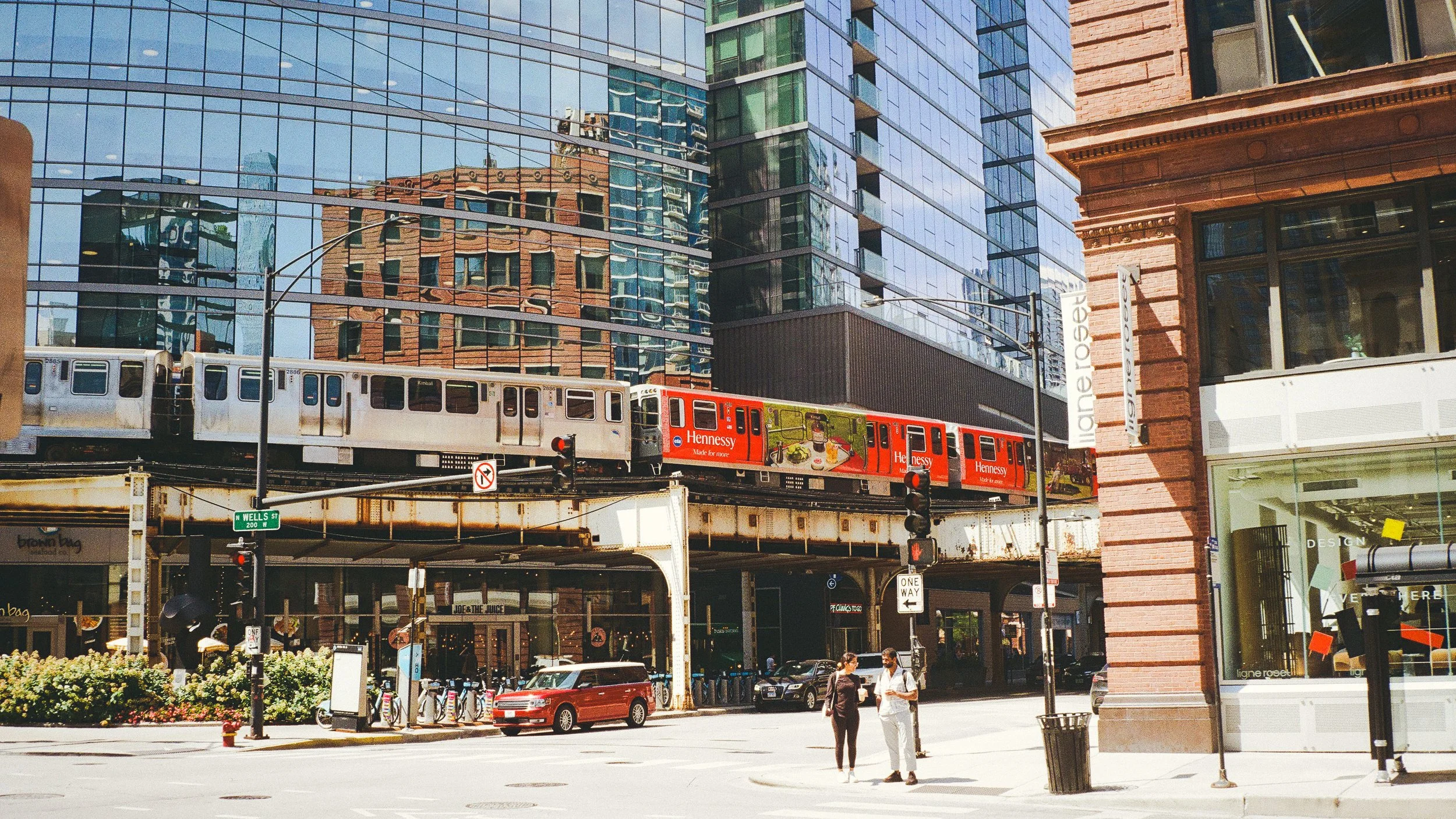 Urban street scene with elevated train on tracks, modern glass buildings, red brick building on the right, pedestrians crossing, cars parked on the street.