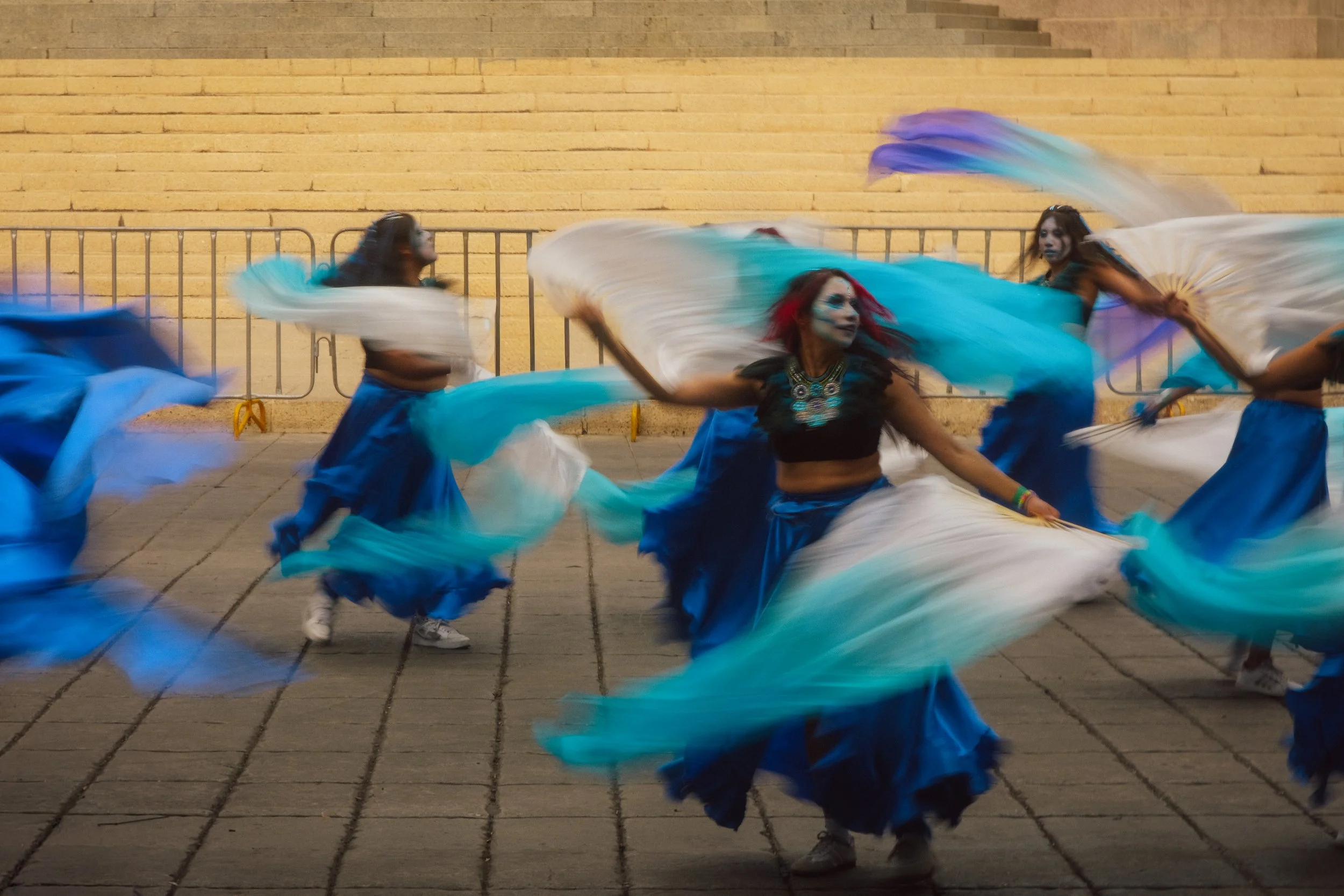 Group of female dancers wearing blue skirts and black tops, performing a traditional dance with large flowing white and blue scarves, in front of a brick wall.