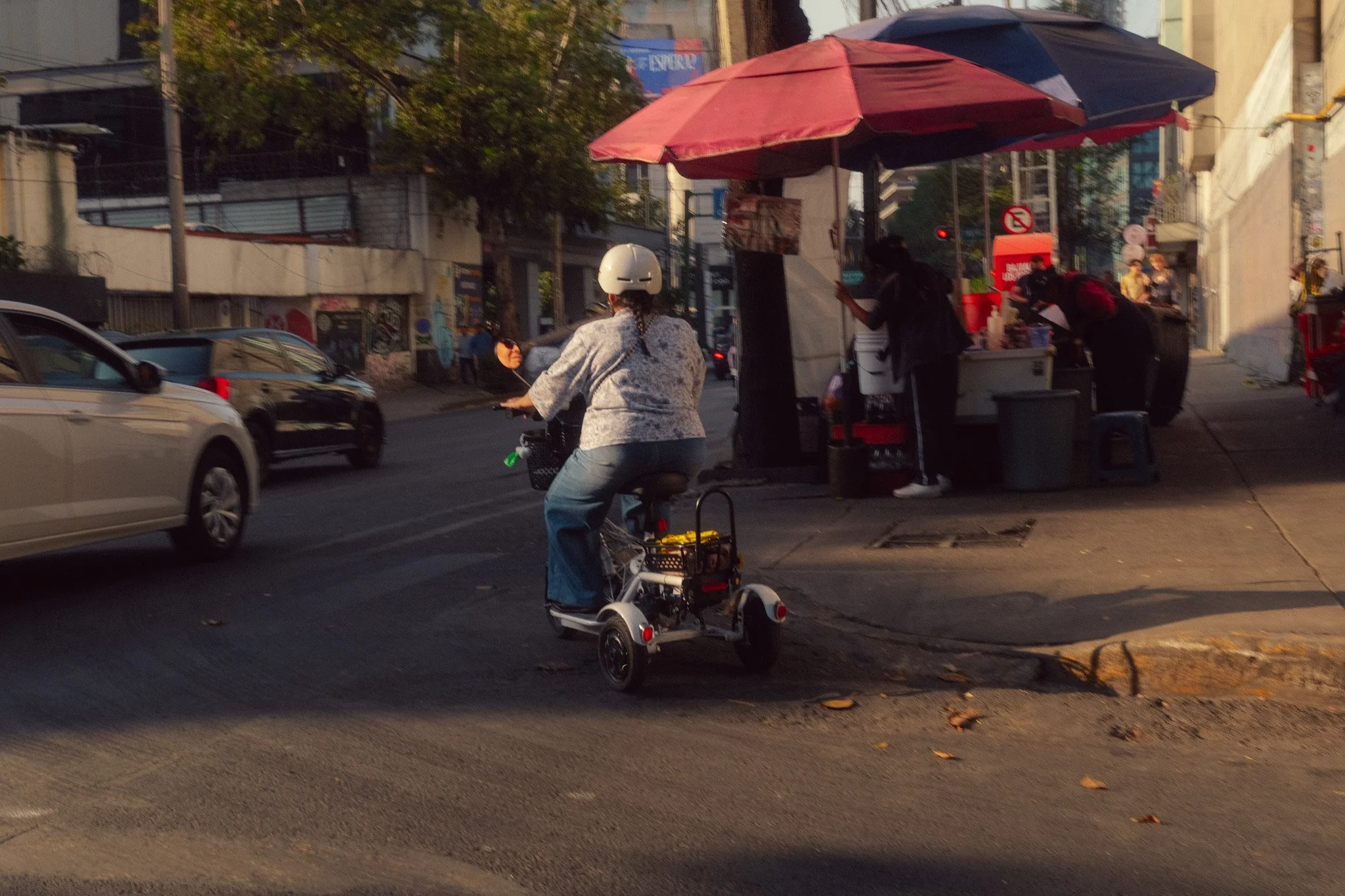 A person riding a mobility scooter on a city street near a small street food stand with red and black umbrellas, and several people working at the stand.