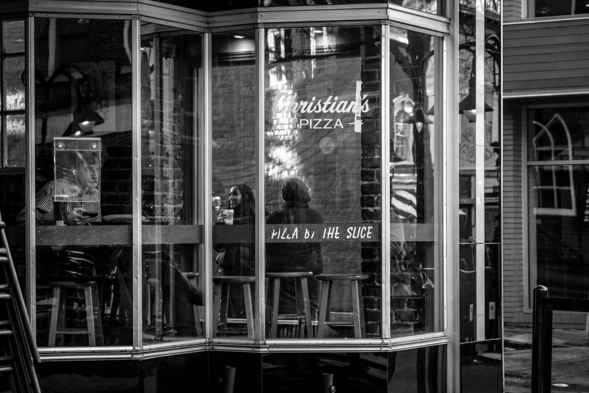 People inside a pizza restaurant named Christian's Pizza, sitting and enjoying their food, seen through a glass window with reflections of the street outside.