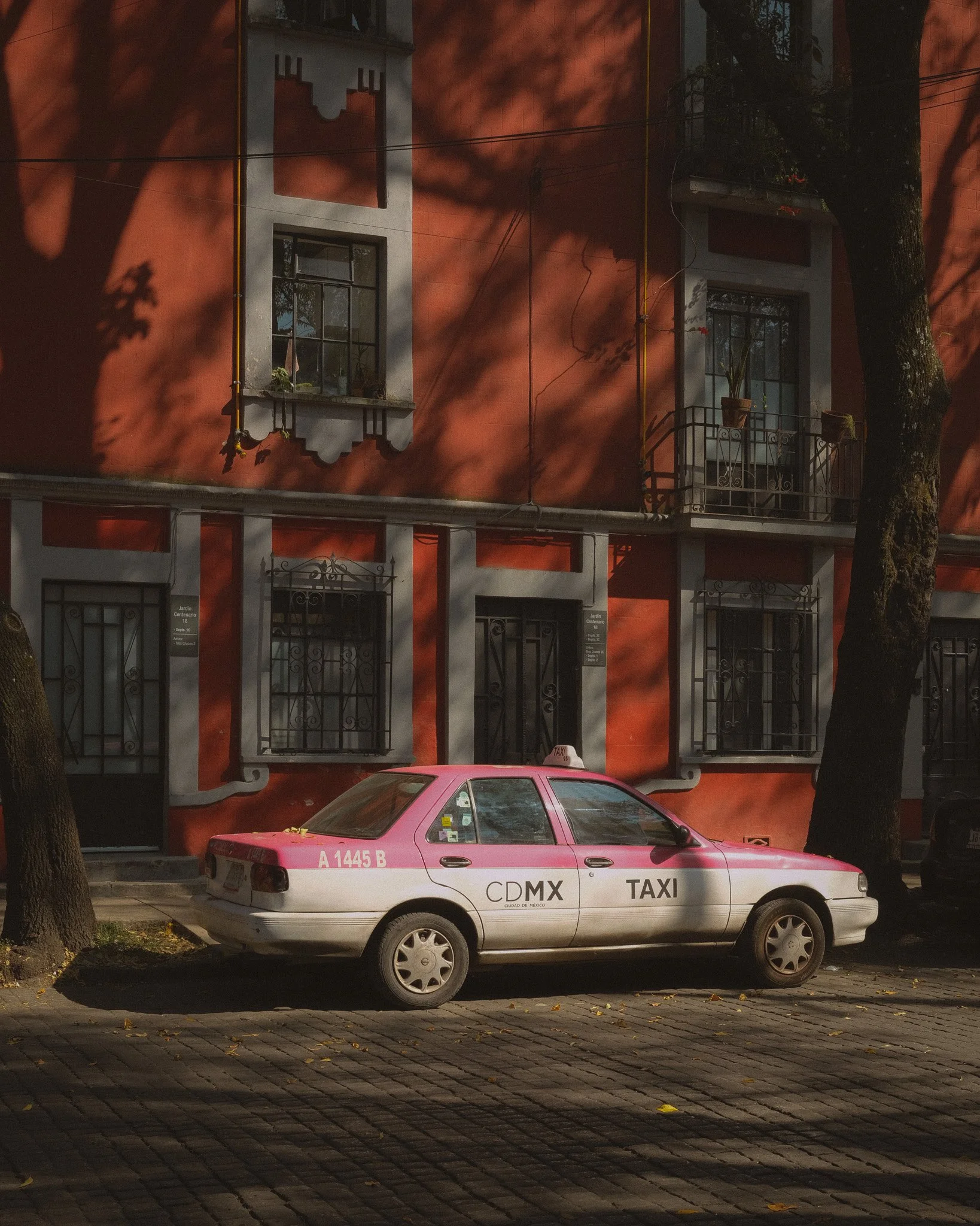 A pink and white taxi parked on a cobblestone street in front of a red building with black wrought iron window grilles and a balcony, with trees shadowing the scene.