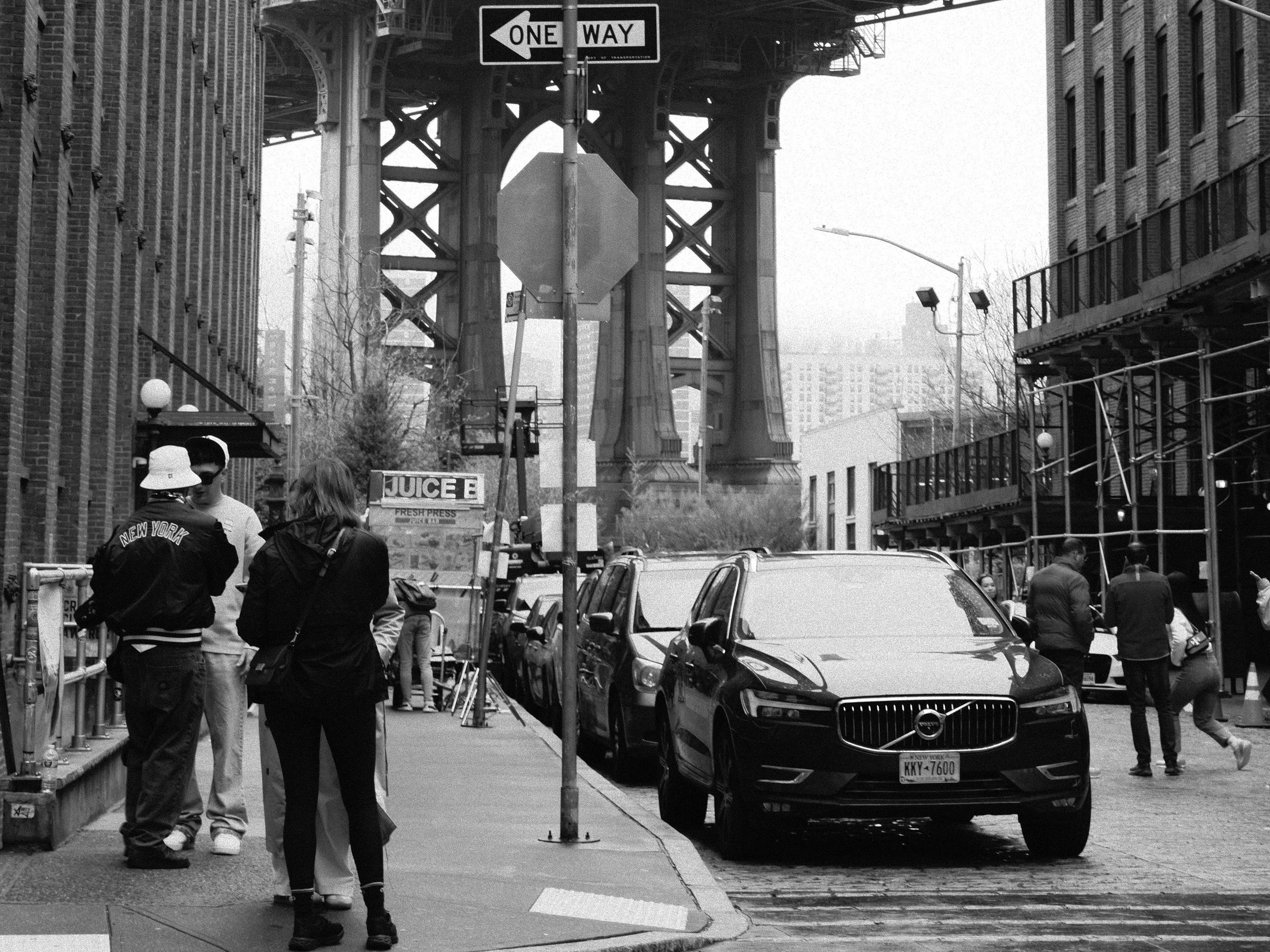Street scene in New York City with people walking, parked cars, brick buildings, a sign for juice, and the Manhattan Bridge in the background.