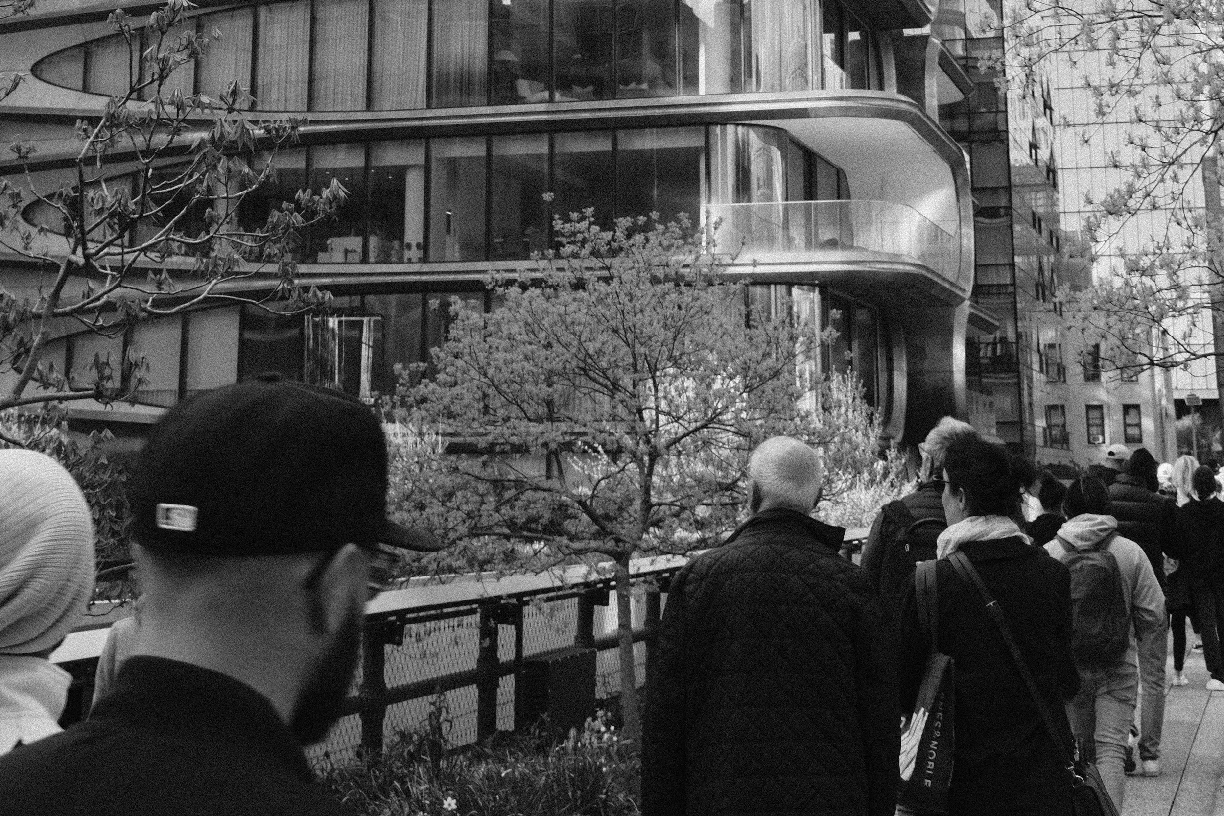 People walking on a city sidewalk near a modern building with curved balconies and glass windows, with trees lining the street.